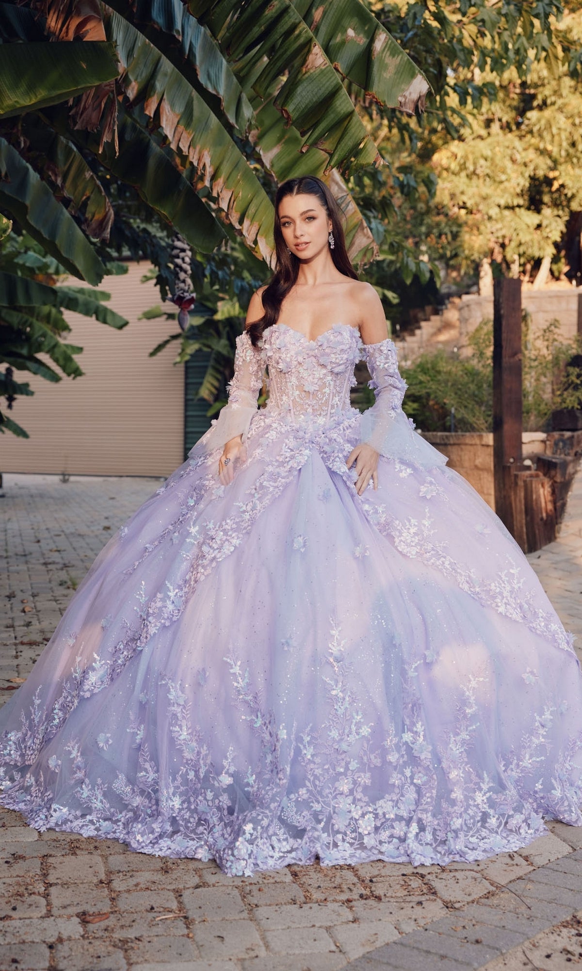 A woman stands outdoors in the Quinceanera Dress JT1459J by Juliet, an off-the-shoulder lilac ball gown with floral embroidery, surrounded by greenery and sunlight.