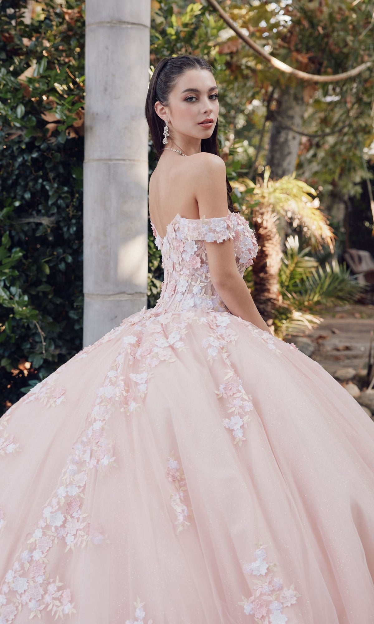 A woman in the Quinceanera Dress By Juliet JT1460J—an off-the-shoulder, blush pink ball gown with floral embellishments—stands outdoors near greenery, looking over her shoulder.