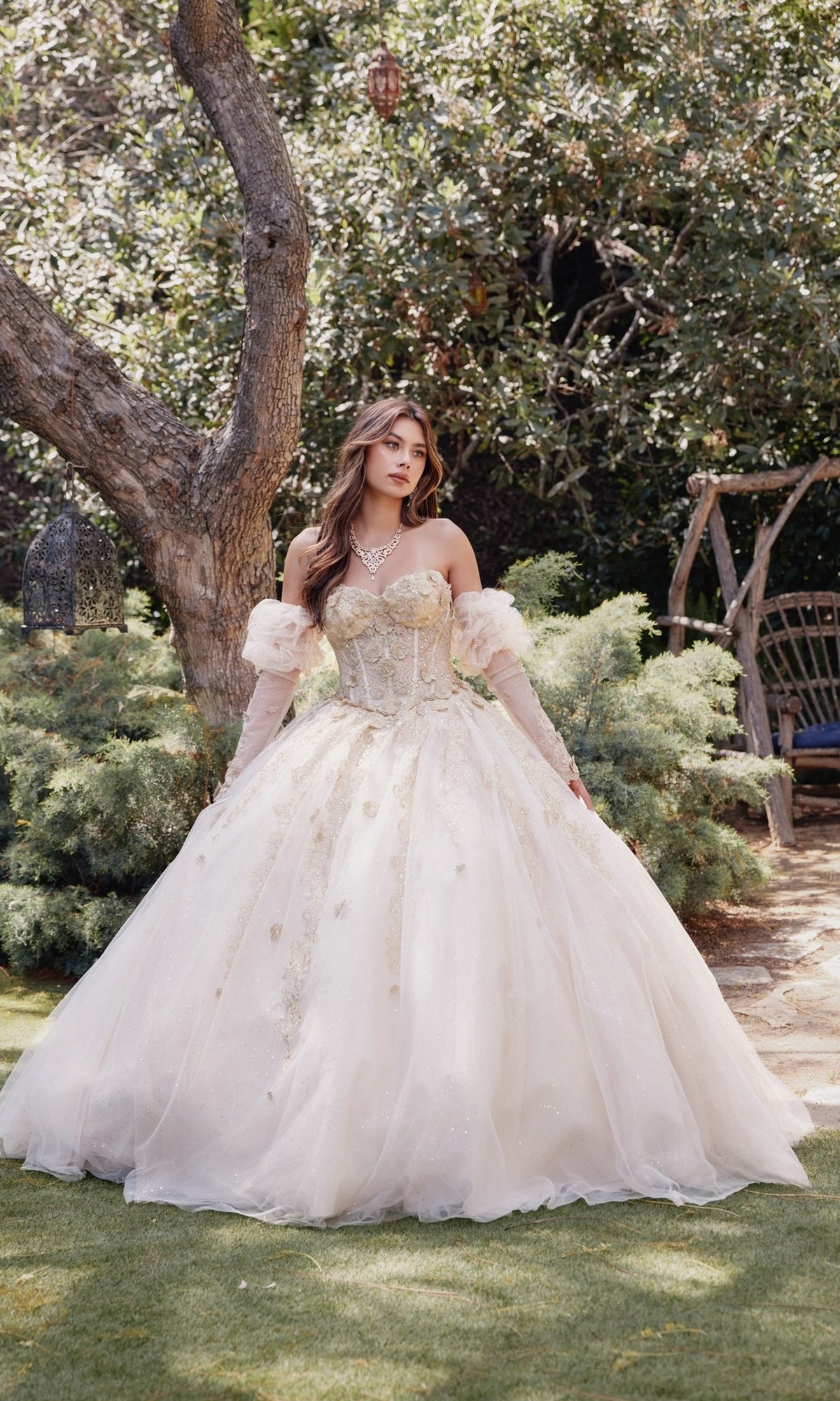 A woman stands outdoors in the Strapless Quinceanera Dress: Juliet JT1465H, featuring a nude hue, full skirt, corset bodice, and long gloves amid trees and lush garden foliage.