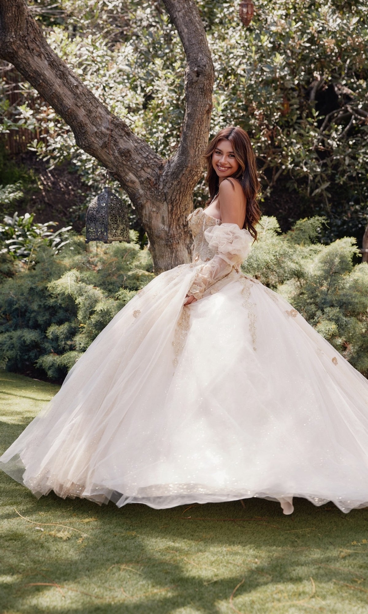 A woman wearing the Strapless Quinceanera Dress: Juliet JT1465H stands on grass outdoors, smiling over her shoulder near a tree and lush greenery.
