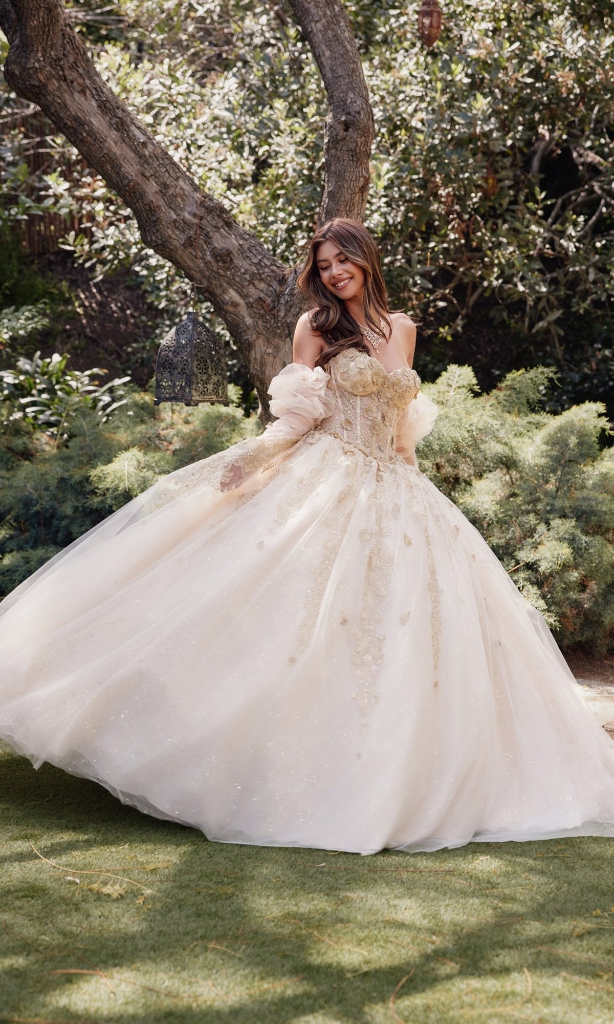 A woman, smiling and looking to the side, stands outdoors on grass in the Strapless Quinceanera Dress: Juliet JT1465H with a voluminous cream skirt, surrounded by trees and greenery.