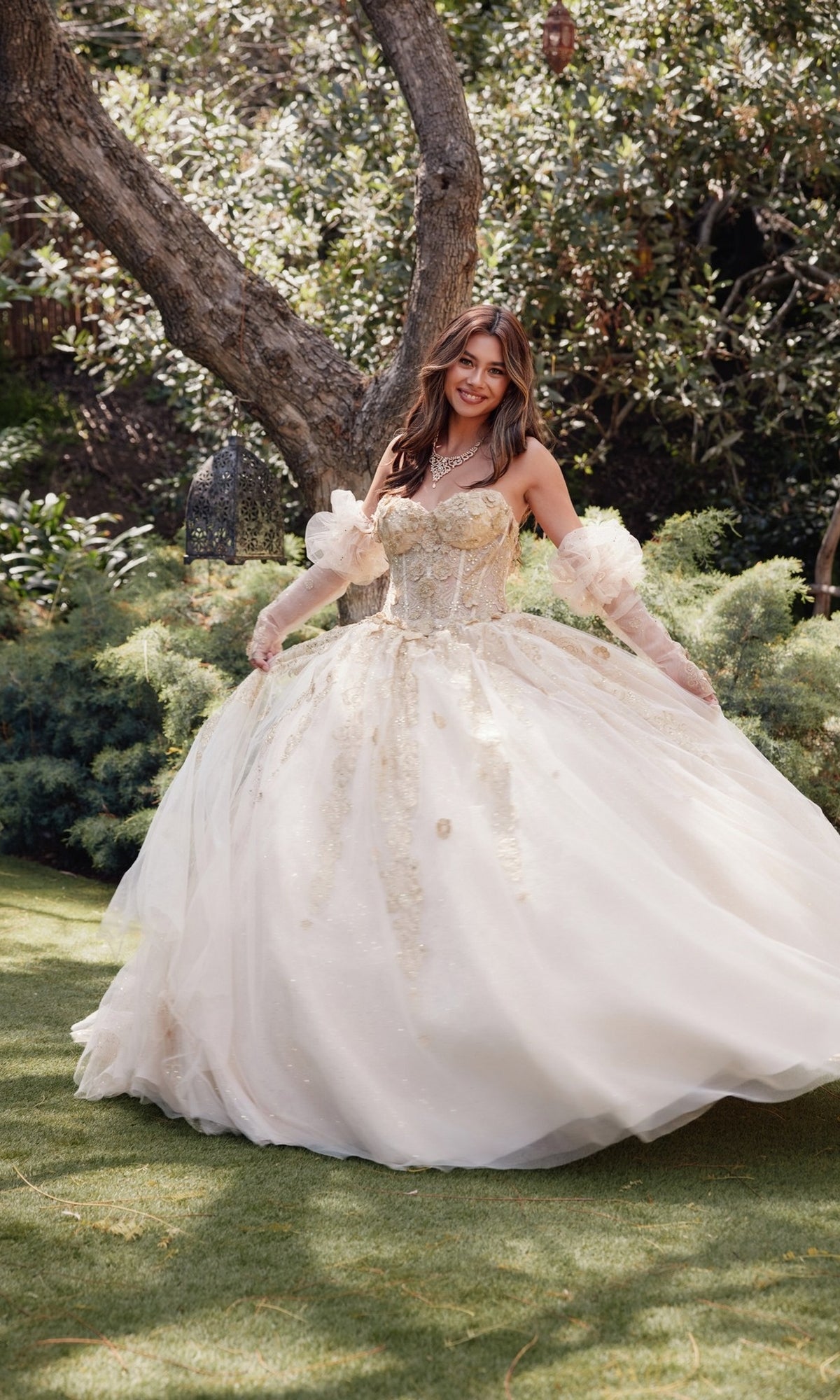 A woman in the Juliet JT1465H Strapless Quinceanera Dress with a corset bodice ball gown stands on grass among greenery and trees, smiling at the camera.