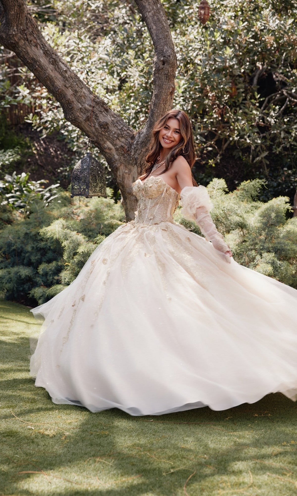A woman wearing the Strapless Quinceanera Dress: Juliet JT1465H stands on grass near a tree, smiling and holding her gown as she twirls outdoors.