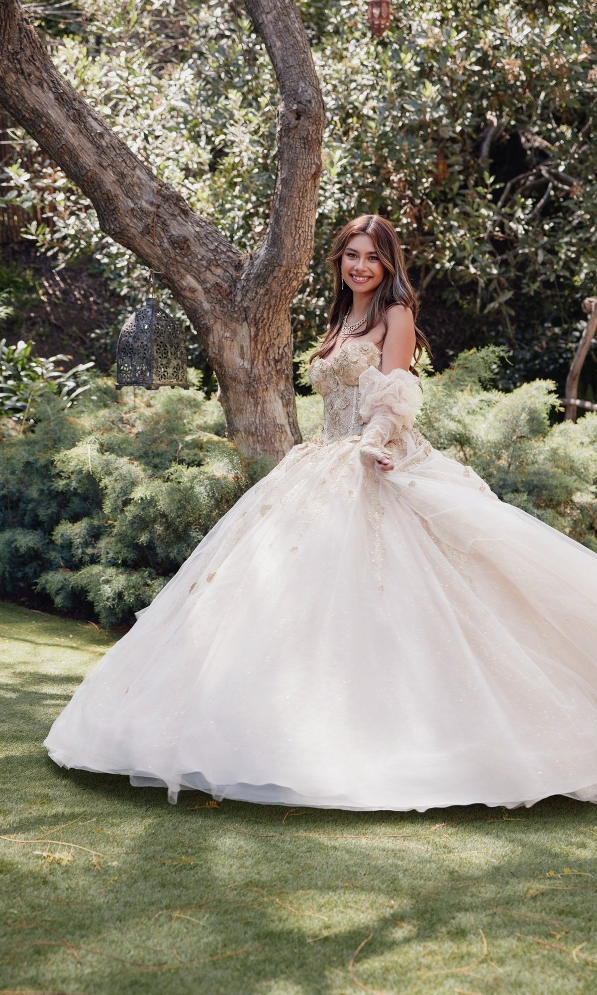 A woman in the Strapless Quinceanera Dress: Juliet JT1465H, featuring a nude corset bodice, stands smiling on grass outdoors with trees and greenery in the background.