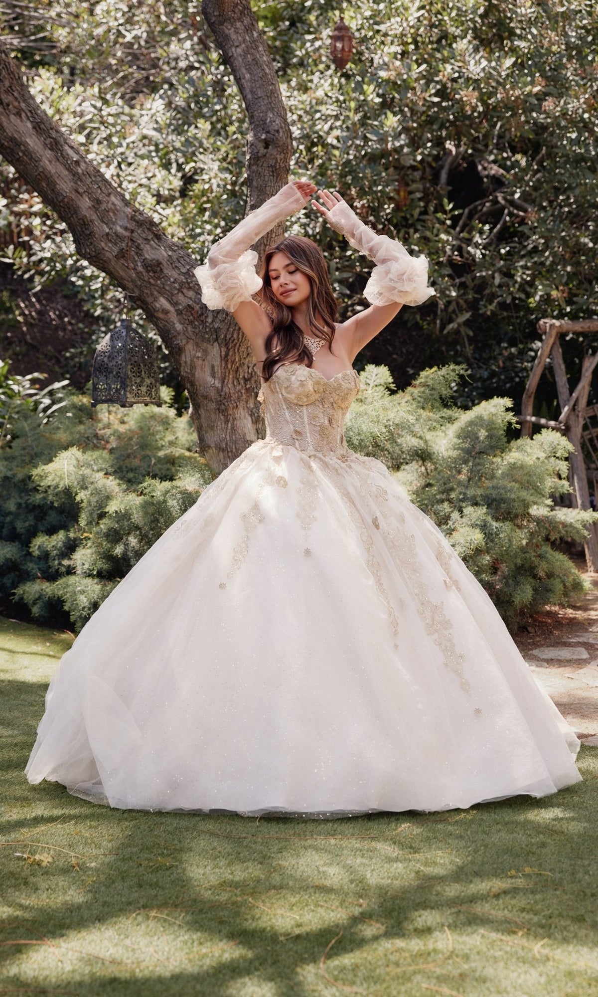 A woman in the Strapless Quinceanera Dress: Juliet JT1465H poses with arms raised in a garden, surrounded by trees and greenery.