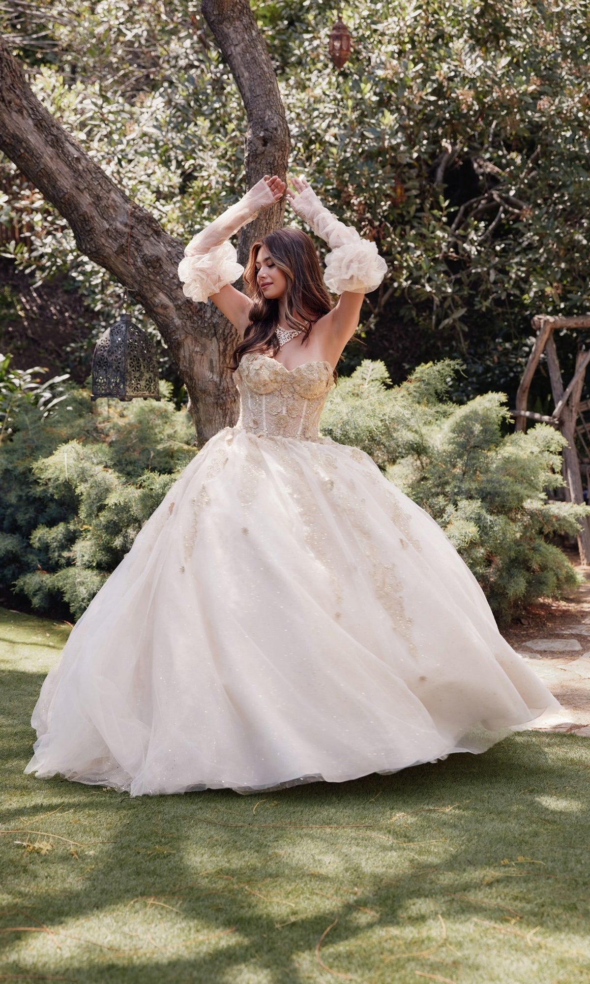 Wearing the Strapless Quinceanera Dress: Juliet JT1465H, a woman stands outdoors on grass amid trees and greenery, her full skirt and sheer gloves completing the elegant look.