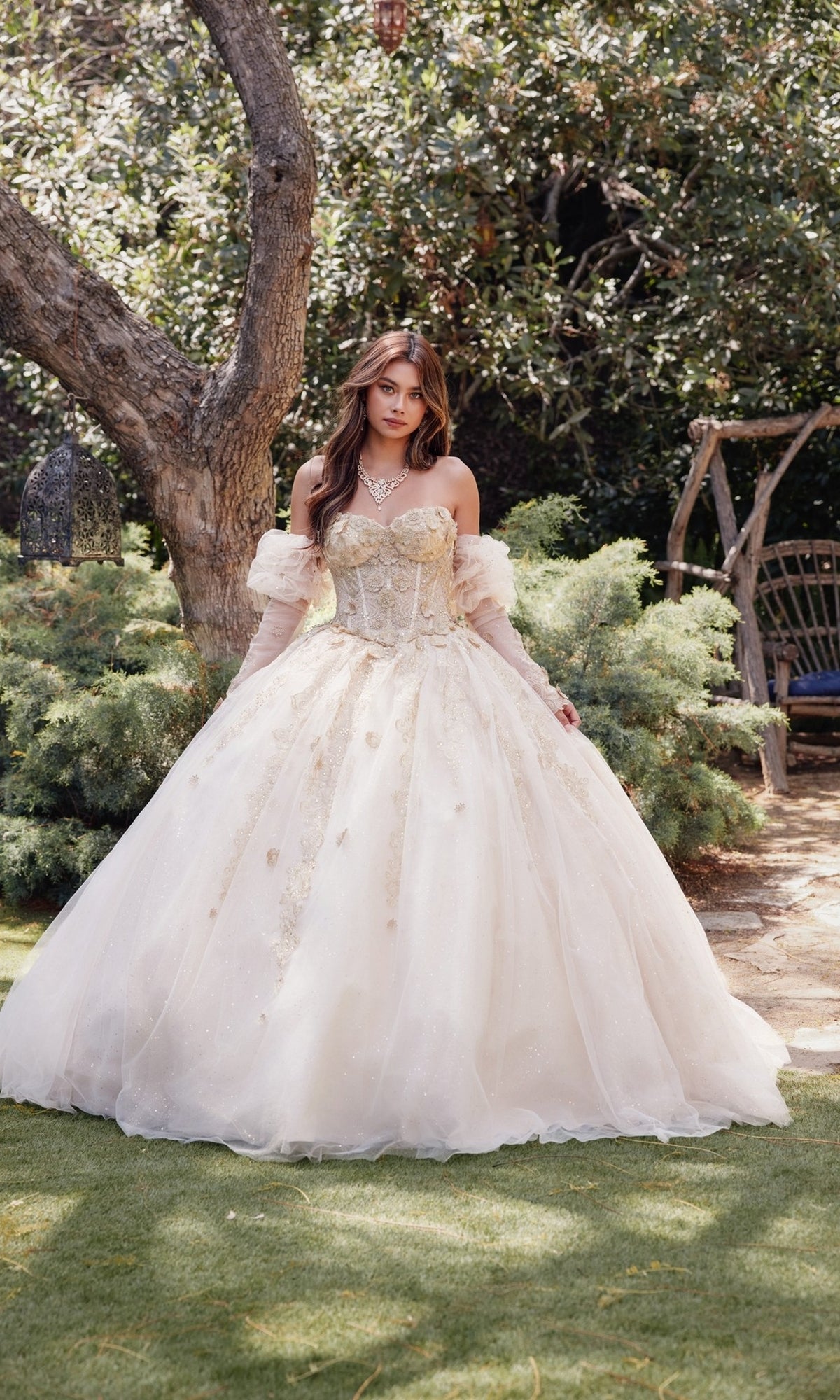 A woman stands outdoors in the Strapless Quinceanera Dress: Juliet JT1465H, featuring a nude-colored, ornate corset bodice and full skirt, surrounded by trees and greenery.
