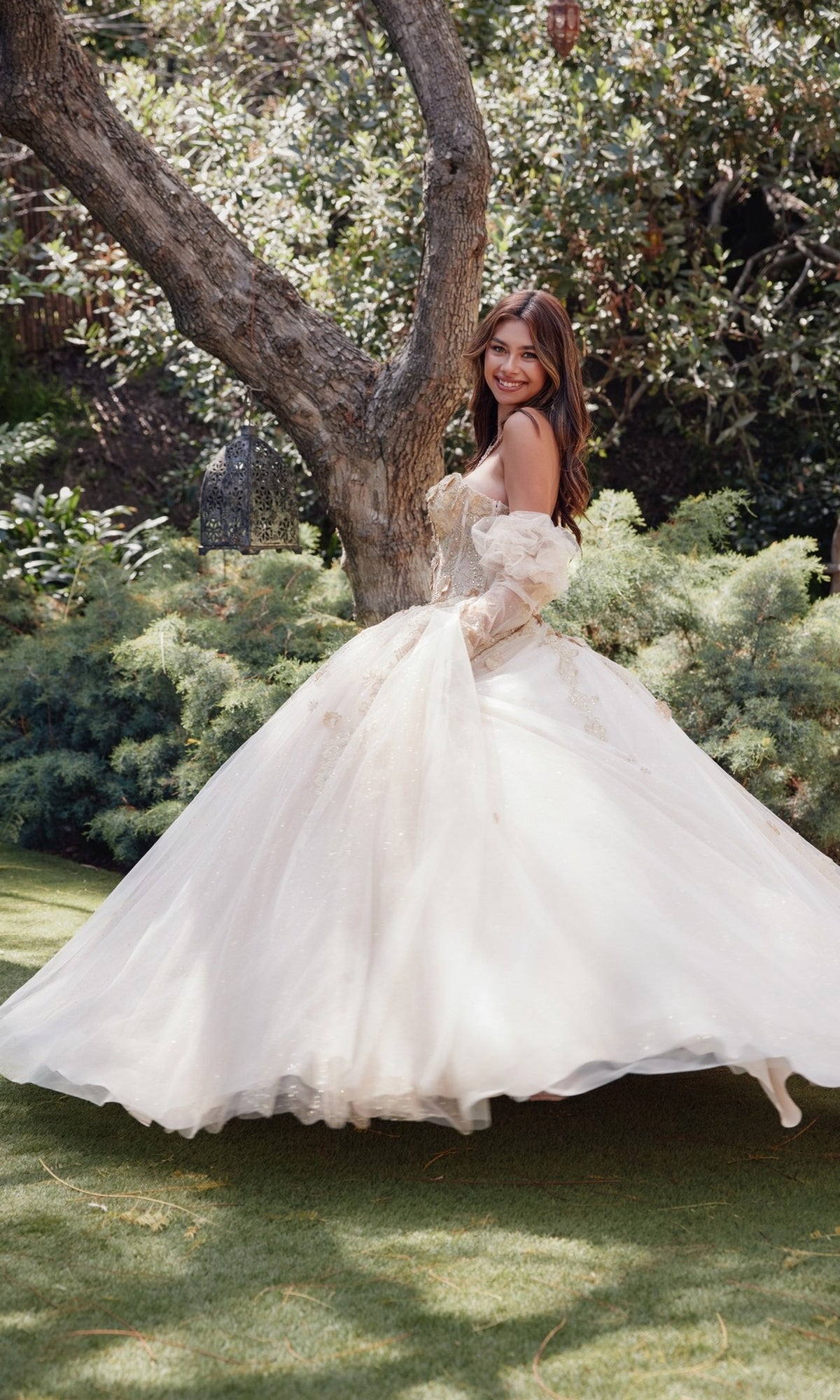 A woman in the Strapless Quinceanera Dress: Juliet JT1465H stands outdoors on grass, smiling and glancing back, surrounded by trees and lush greenery in her corset bodice ball gown with a full skirt.