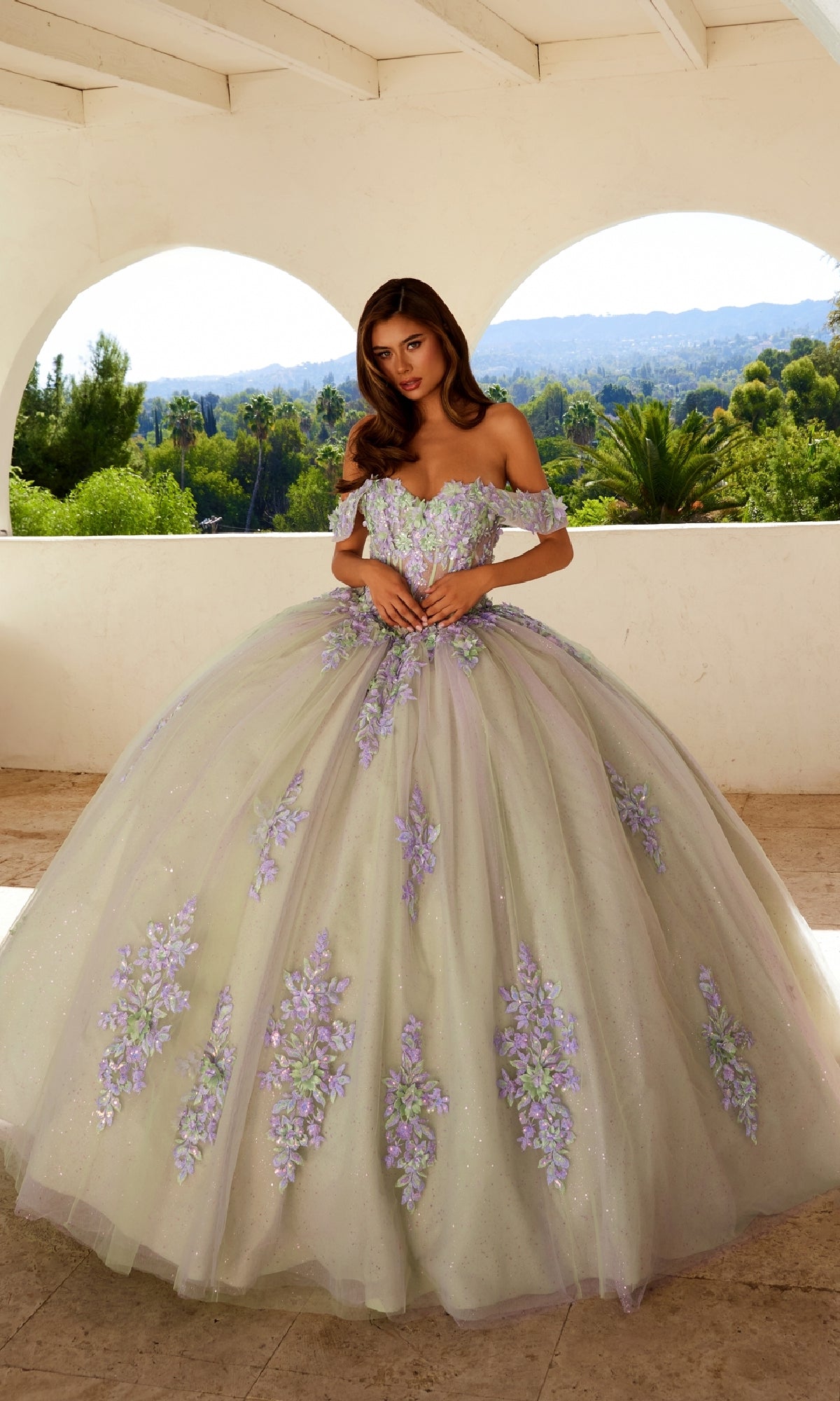 A woman stands on a terrace in the Quinceanera Dress JT1479J By Juliet, which features lilac floral embroidery and a glitter tulle ball gown skirt, surrounded by greenery and sunlight.