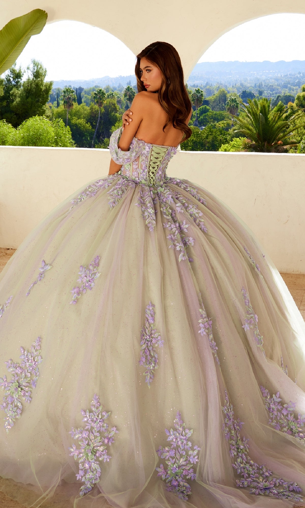 A woman faces away outdoors under a greenery-adorned arched veranda, wearing the Quinceanera Dress JT1479J By Juliet—a large pastel gown with floral embroidery.
