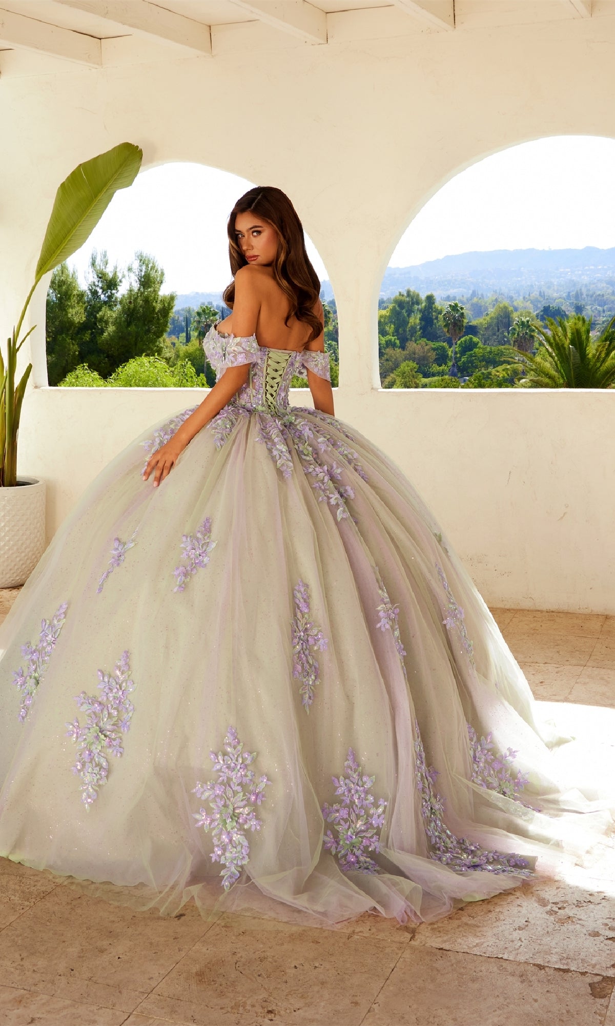 A young woman wears the Quinceanera Dress JT1479J By Juliet, an off-the-shoulder pastel gown with purple floral embroidery, glitter tulle accents, and a full skirt, as she stands on a terrace looking over her shoulder.