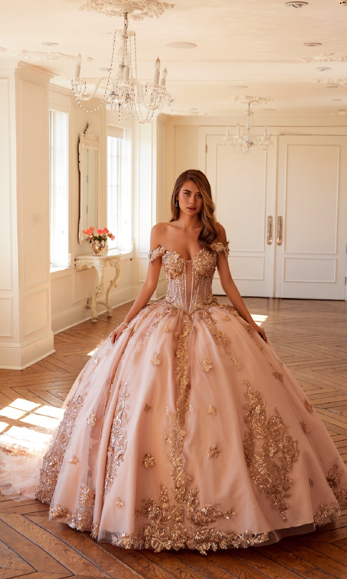 A woman stands in a well-lit room wearing the Quinceanera Dress JT1485J By Juliet, an ornate, off-the-shoulder gown in pink with gold embroidery and a full skirt.