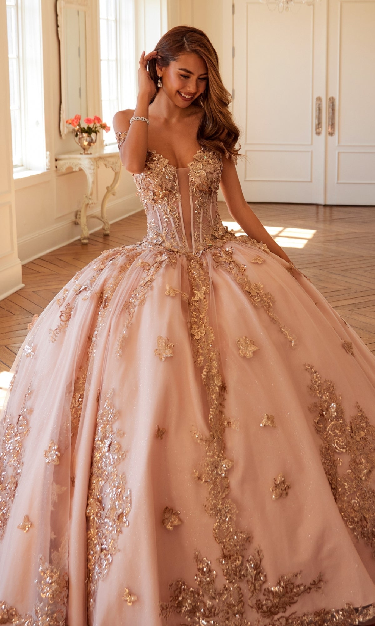 A woman wears the Quinceanera Dress JT1485J By Juliet, an off-the-shoulder blush pink ball gown with gold floral embellishments, as she stands in a bright room with wooden floors and white walls.
