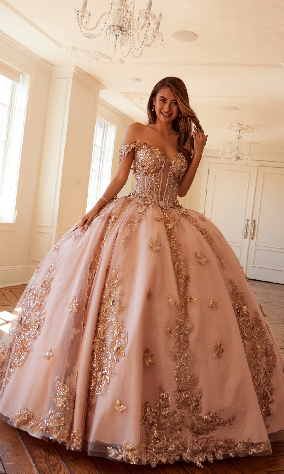 A woman stands in a bright room wearing the elegant Quinceanera Dress JT1485J By Juliet, featuring pink fabric, gold floral appliqués, and off-the-shoulder sleeves.