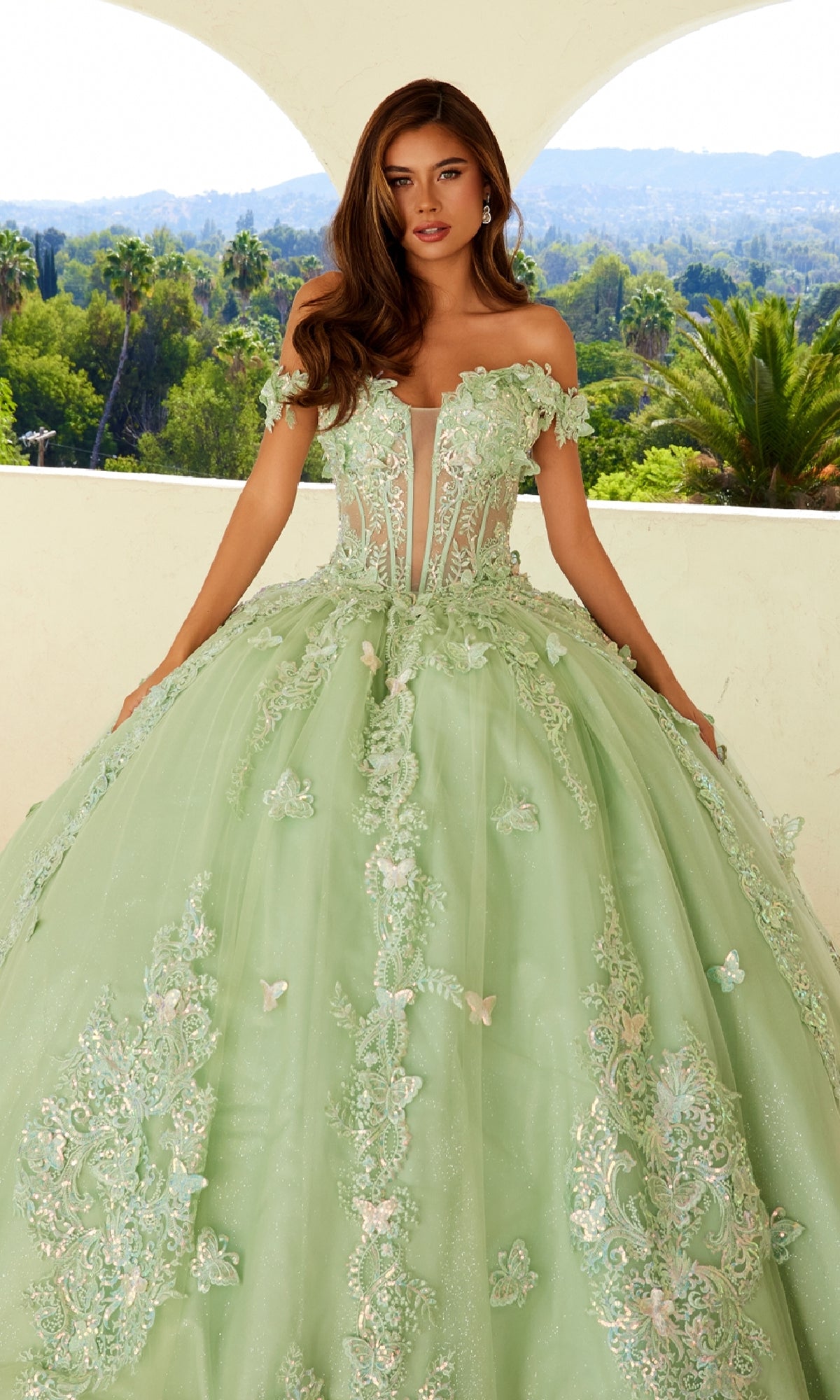 A woman stands on a balcony wearing the Quinceanera Dress JT1485J By Juliet, a light green off-the-shoulder gown with floral lace details and embellishments. Lush greenery and hills are visible in the background.