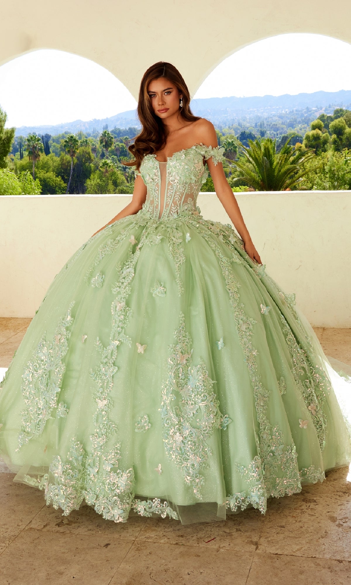 A woman wearing the Quinceanera Dress JT1485J By Juliet, a light green off-the-shoulder gown with floral and butterfly appliqués, stands outdoors before an arched balcony and a lush landscape.