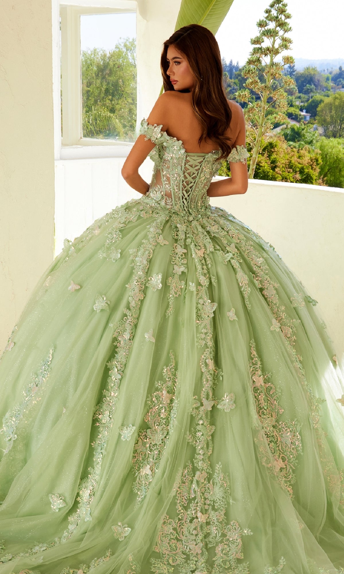 A woman stands on a balcony wearing Juliet's Quinceanera Dress JT1485J in pastel green, featuring an off-the-shoulder design with floral lace appliqué and a full tulle skirt.