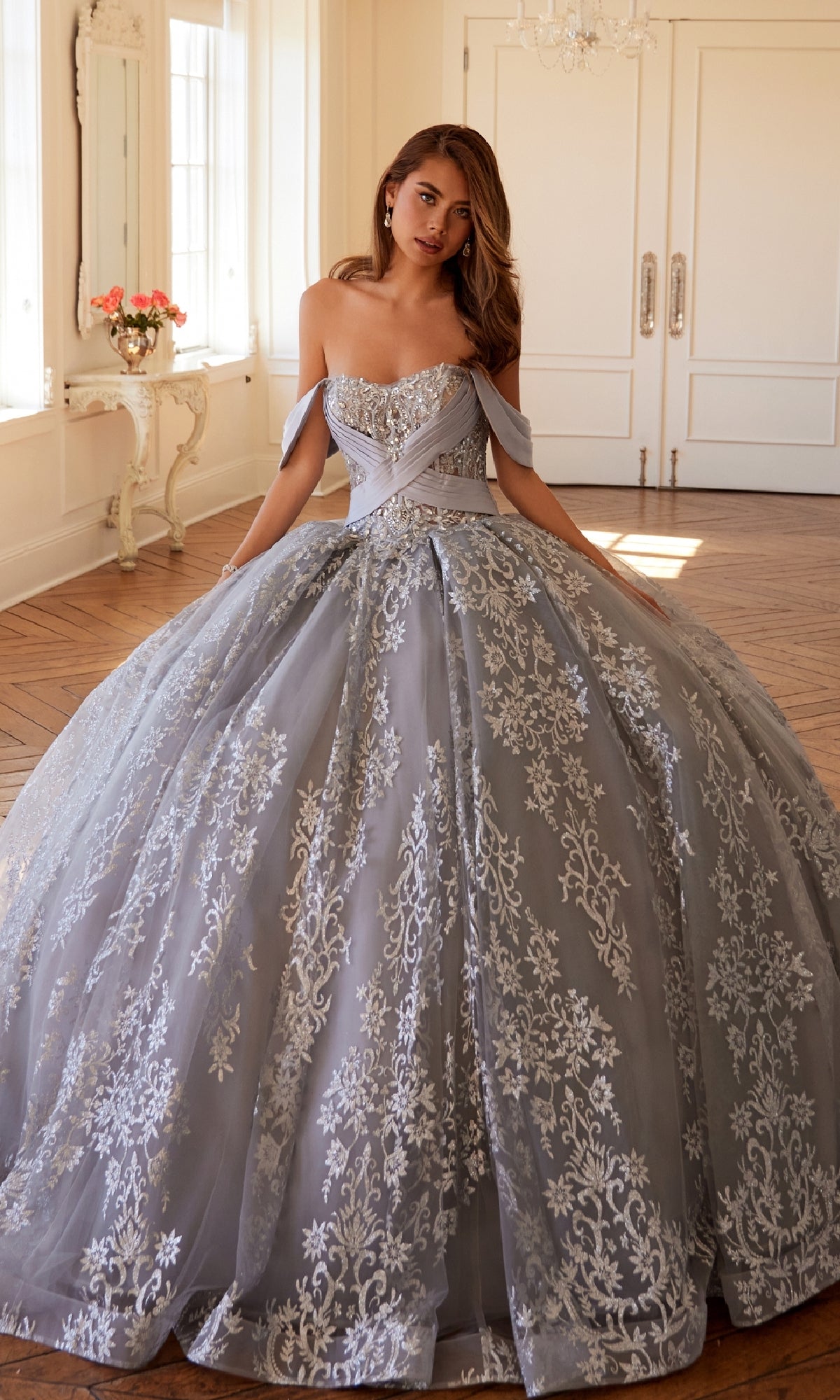 A woman stands indoors wearing the Quinceanera Dress JT1488H By Juliet, featuring a light blue hue, silver embroidery, an off-the-shoulder corset bodice, and a flowing ball gown skirt.