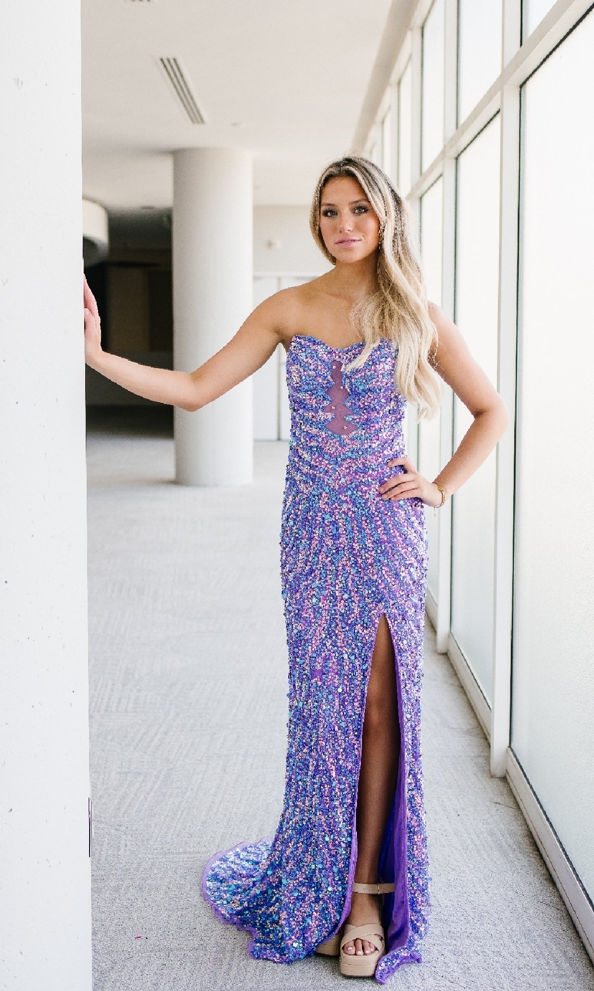 A woman in the Aleta 1504 long purple prom dress with sparkling sequins and a thigh-high slit stands indoors by a large window, resting her arm on a white column.