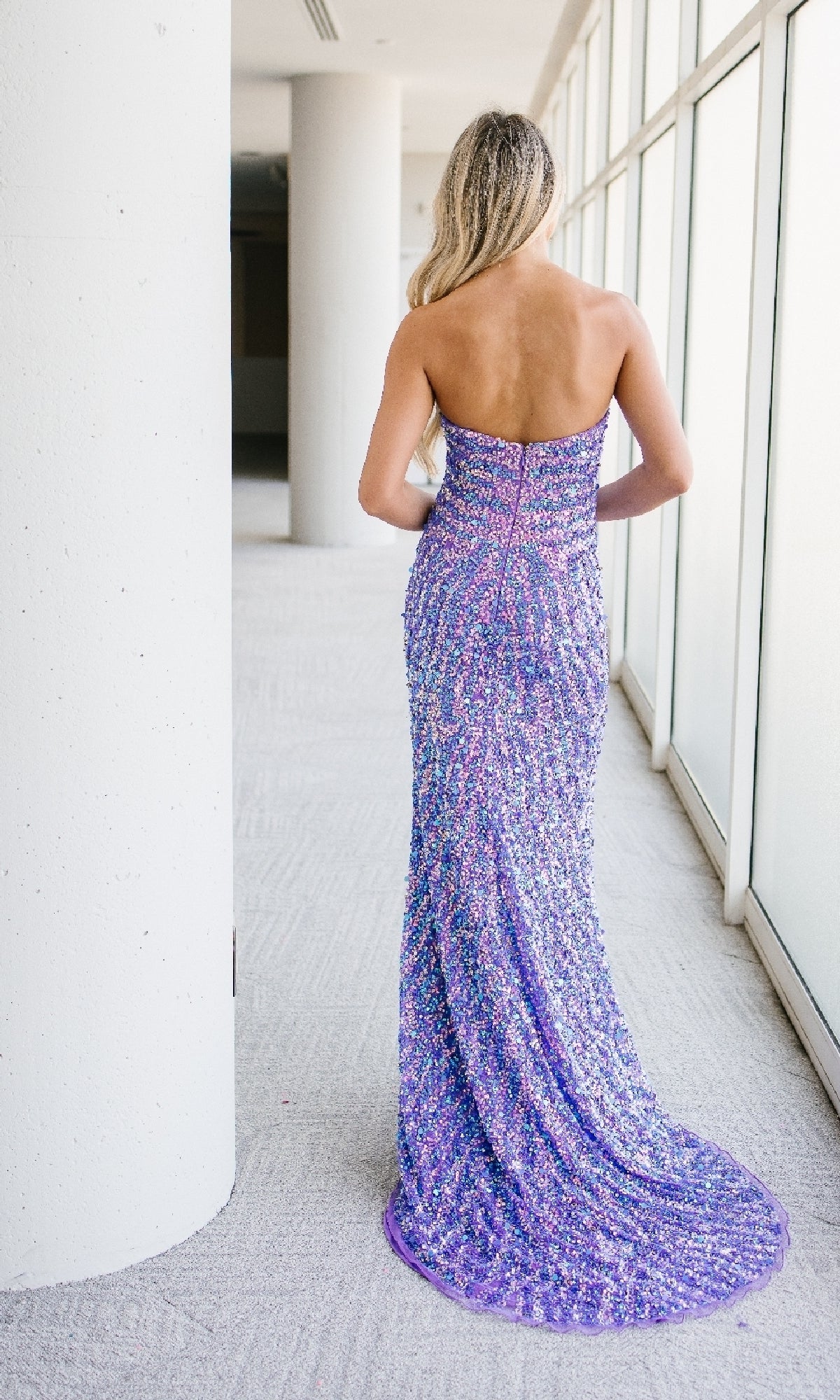A woman with long blonde hair stands facing away in a bright hallway, wearing the Aleta 1504 Long Purple Prom Dress adorned with sparkling sequins.