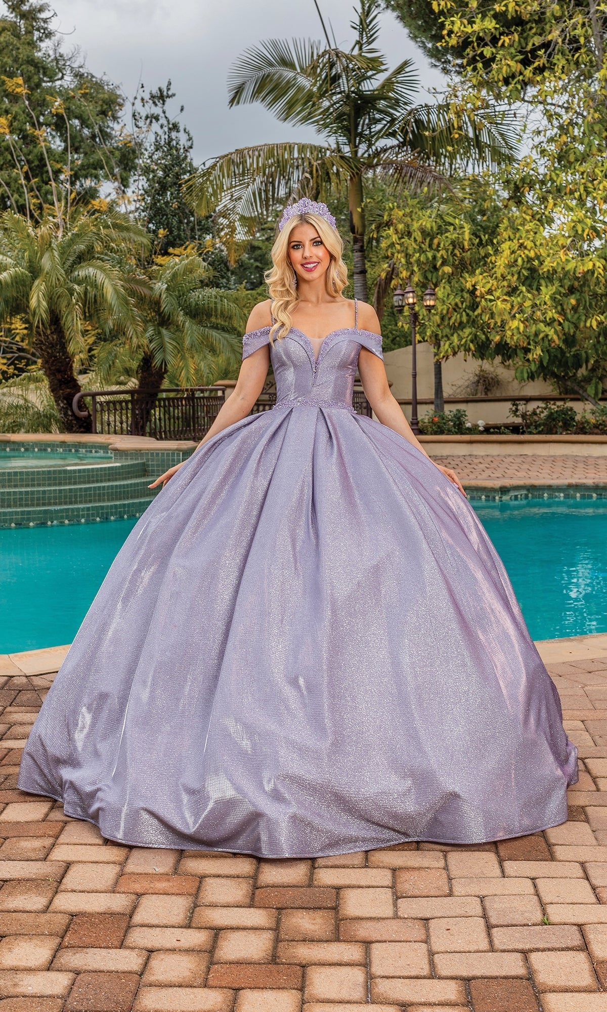 A woman in the Quinceanera Dress 1506 by Dancing Queen, a sparkling lavender off-shoulder ball gown, stands by a pool on a tiled patio with palm trees and lush greenery in the background.
