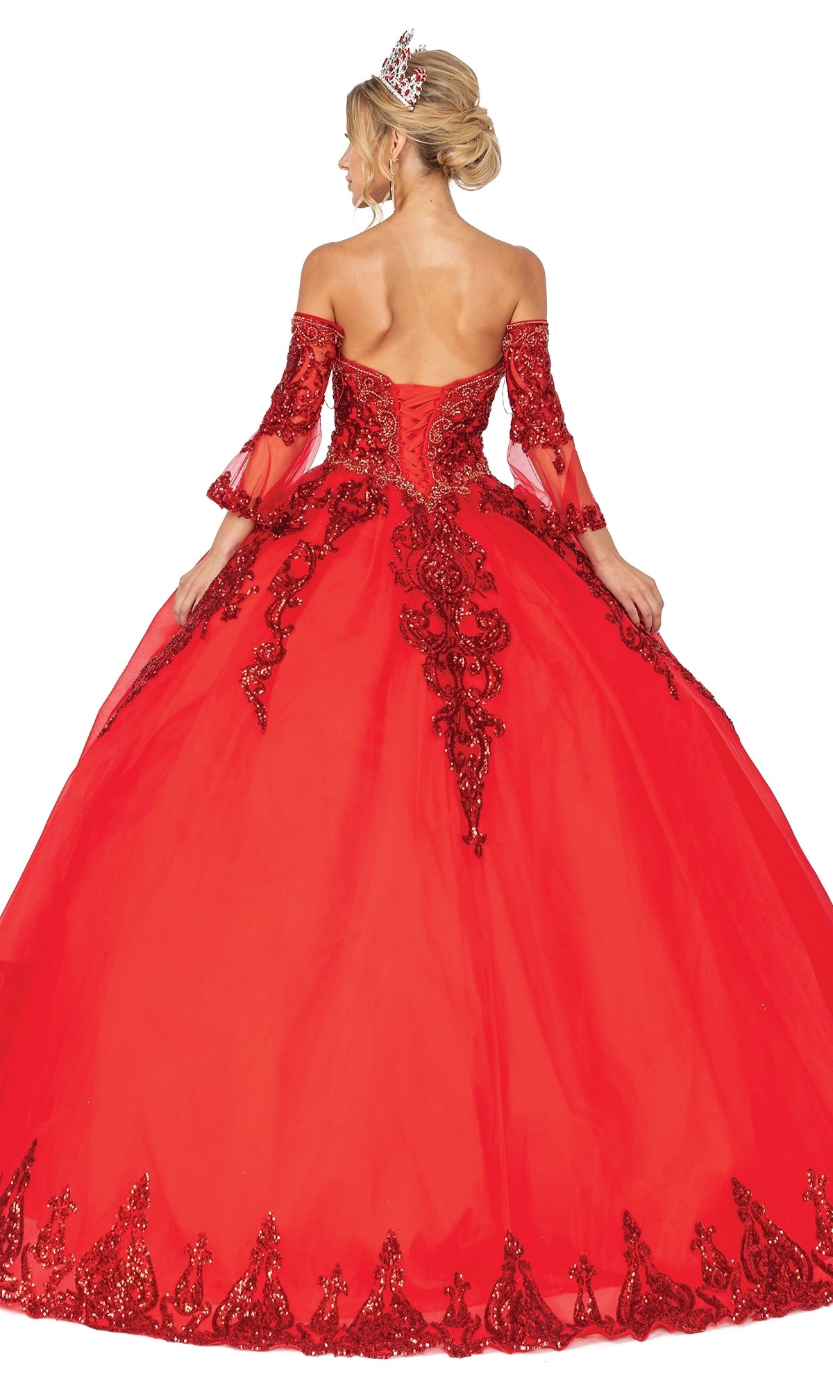 A woman with an updo and tiara wears the Quinceanera Dress 1512 by Dancing Queen, featuring an a-line ball gown silhouette, lace and sequin details, removable sheer sleeves, and her back turned to the camera.
