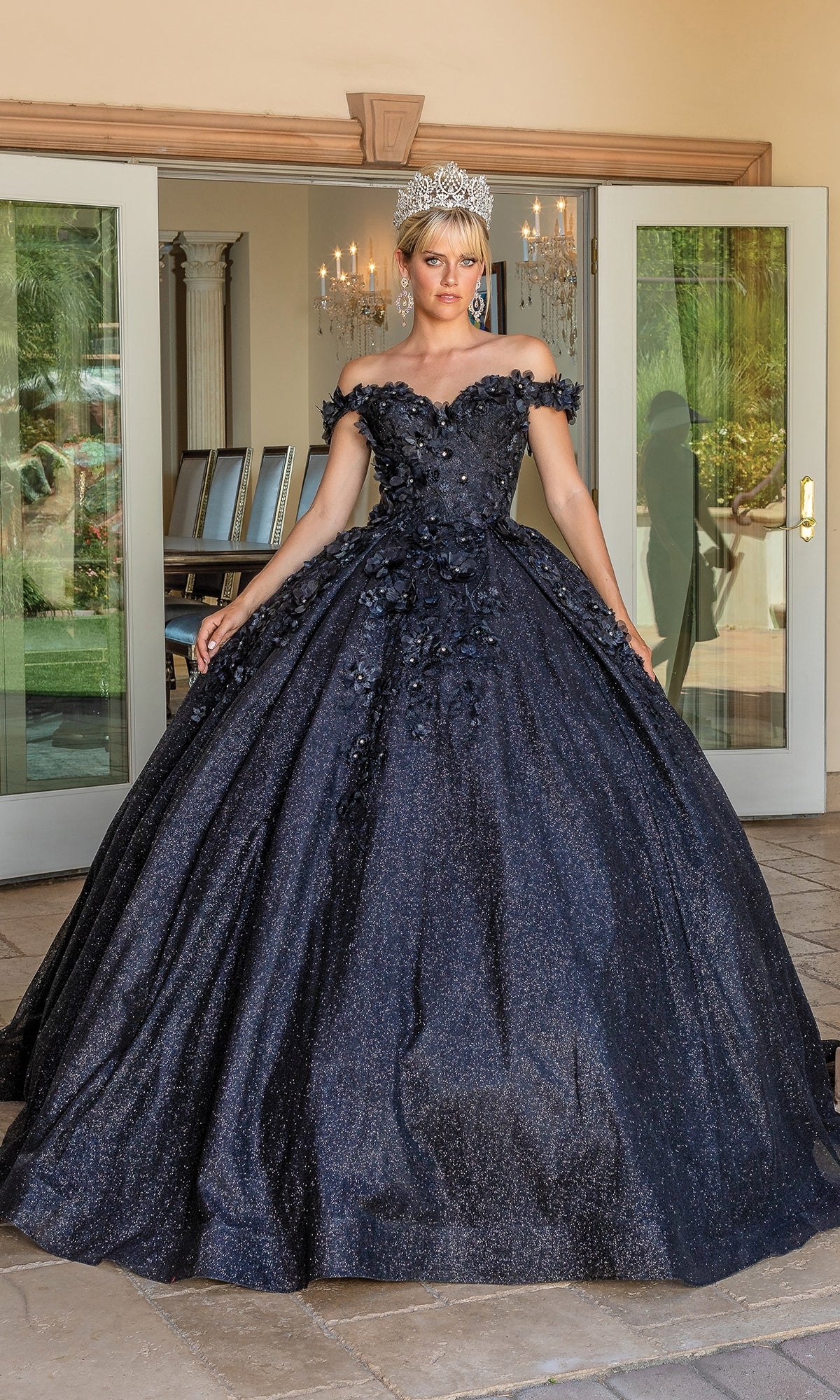 A person stands before glass doors wearing the Quinceanera Dress 1527 by Dancing Queen, a dark blue off-the-shoulder ball gown featuring floral details, sparkling texture, and accessorized with a silver tiara.