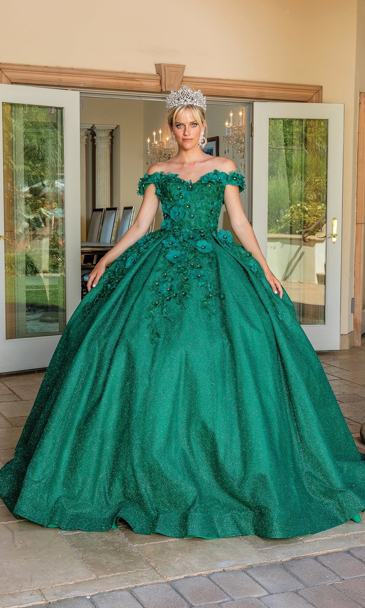 A woman wearing the Quinceanera Dress 1527 by Dancing Queen—a sparkling green ball gown with floral accents and a sweetheart neckline—stands before double glass doors, crowned in silver.