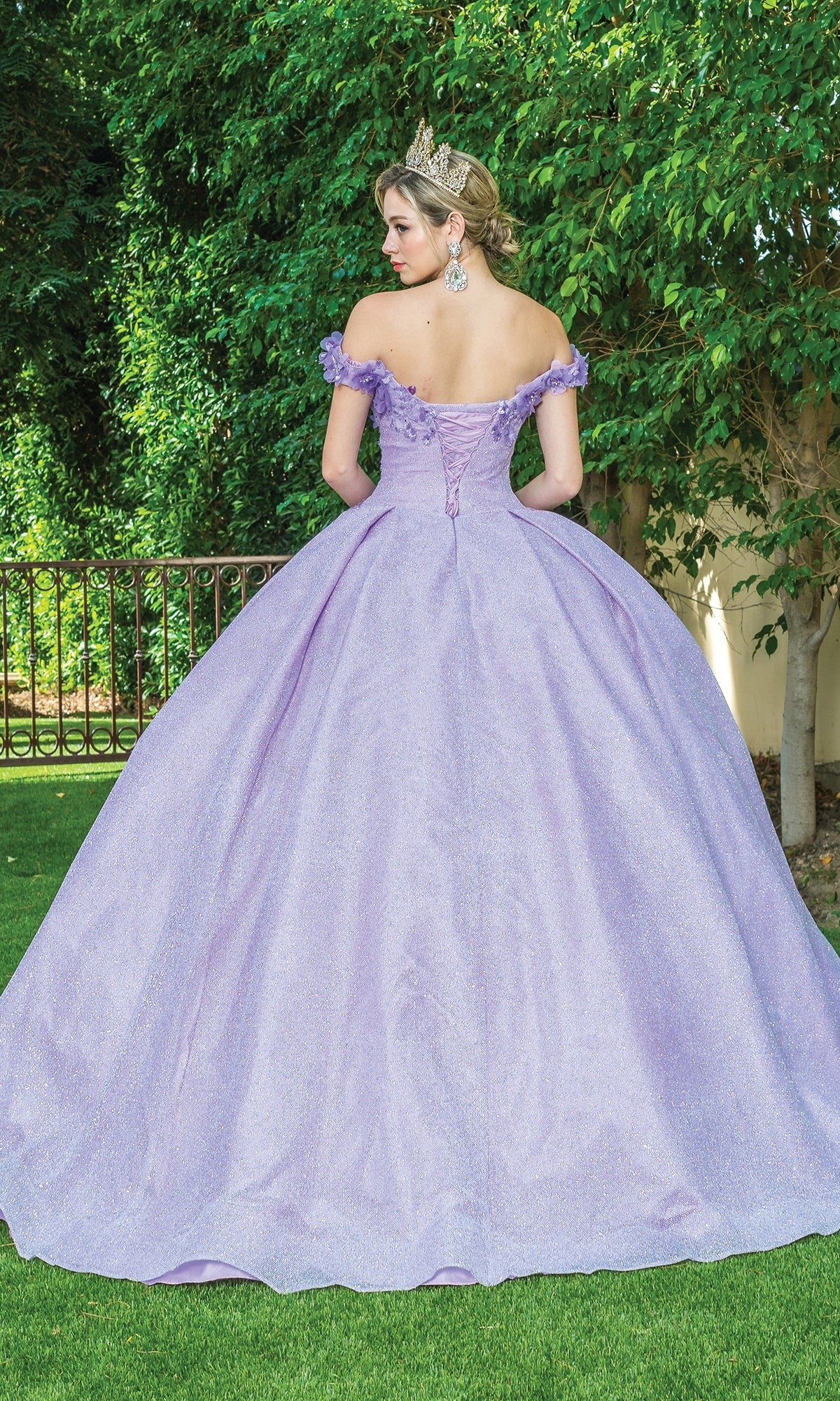 A woman in the Quinceanera Dress 1527 by Dancing Queen, a lavender off-the-shoulder ball gown, wears a crown and stands with her back to the camera on grass, surrounded by trees and a metal fence.
