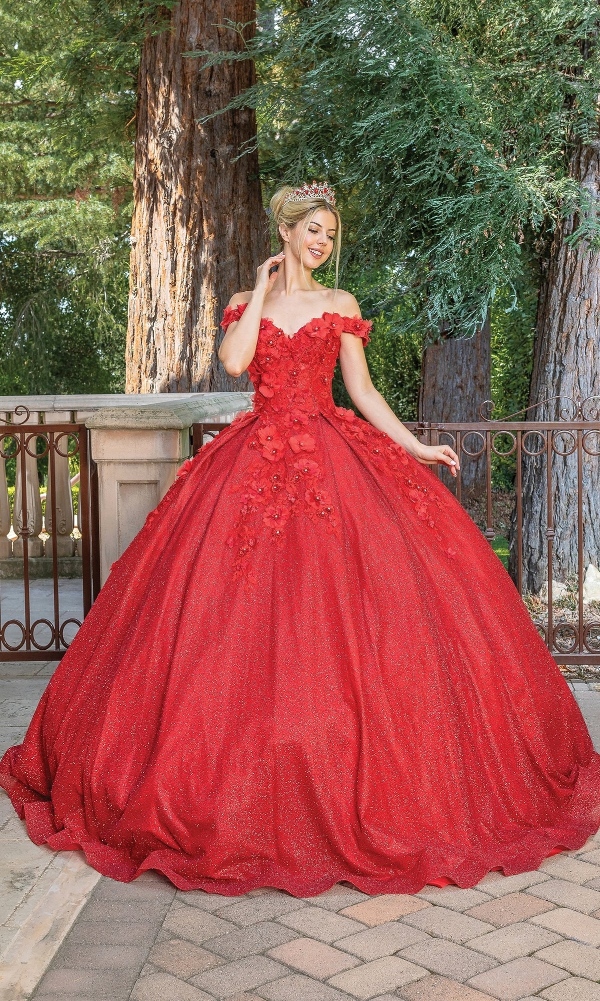 A woman wearing the Quinceanera Dress 1527 by Dancing Queen, a bright red, off-the-shoulder ball gown with floral details and a sweetheart neckline, stands outdoors on a brick path, framed by trees and a wooden railing.