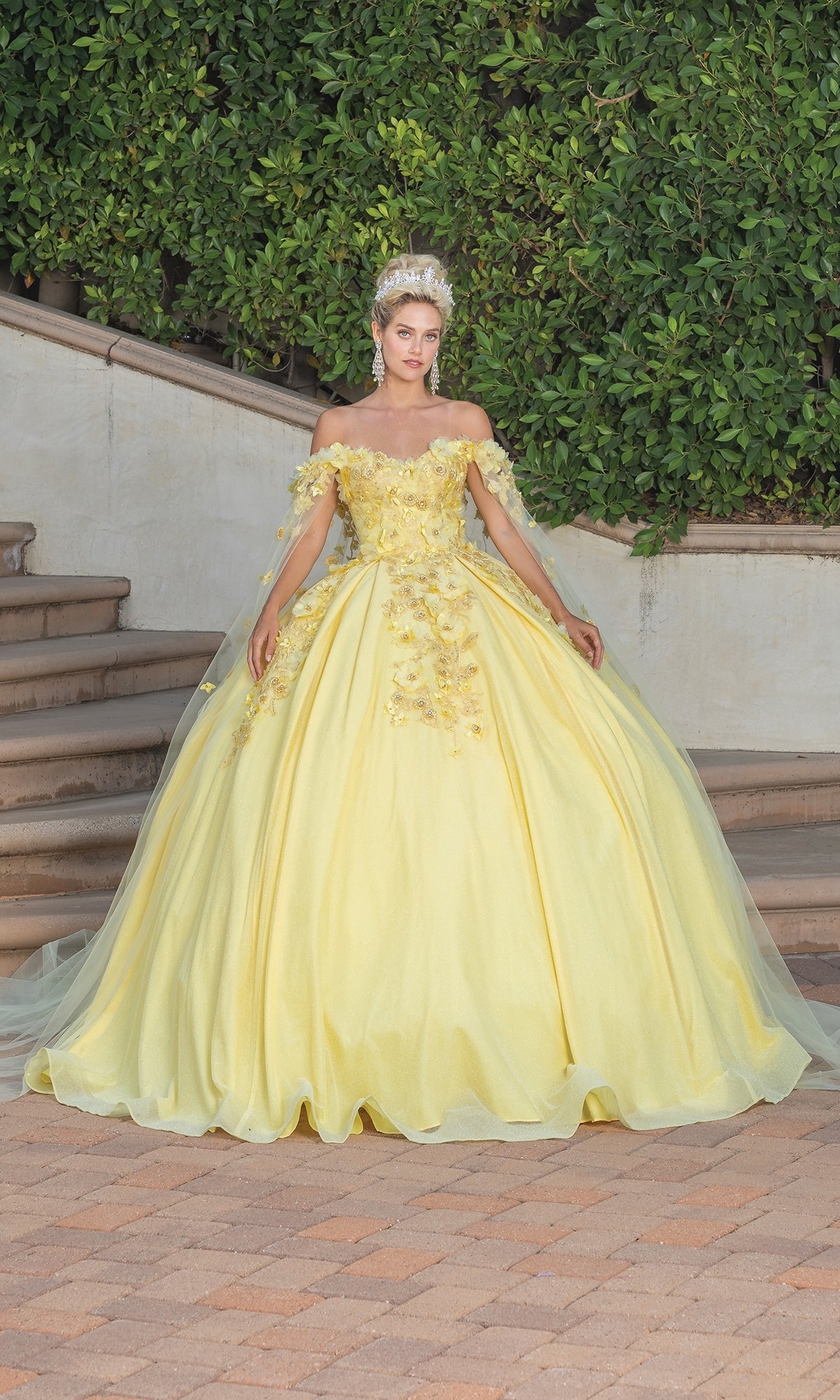 A woman stands on stone steps in lush greenery, wearing the Quinceanera Dress 1527 by Dancing Queen—an ornate ball gown with floral embellishments, a sweetheart neckline, off-the-shoulder sleeves, and a sparkling tiara.