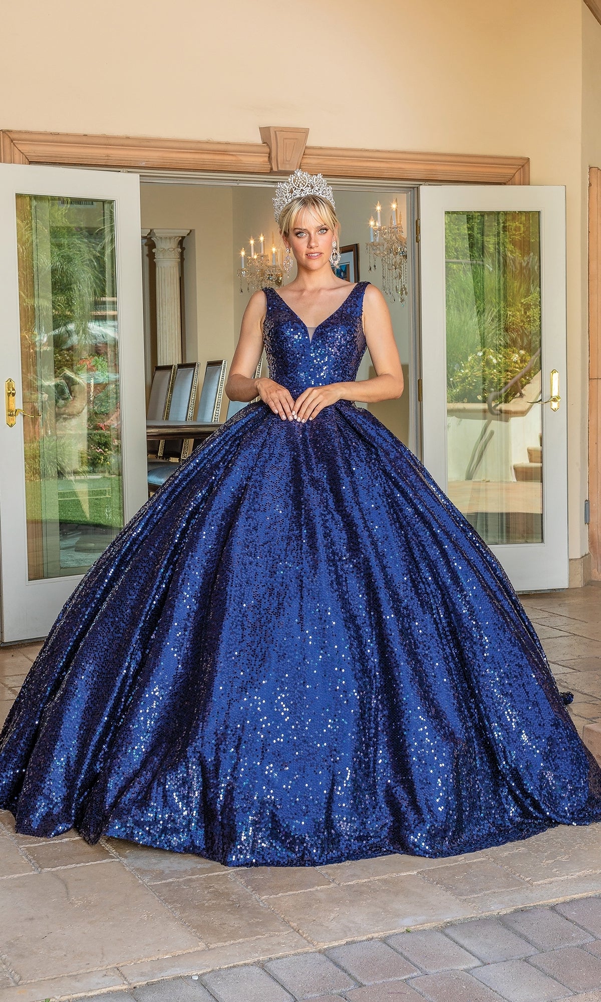 A woman in the Quinceanera Dress 1558 by Dancing Queen and a tiara stands before open double doors, with chandeliers visible inside.