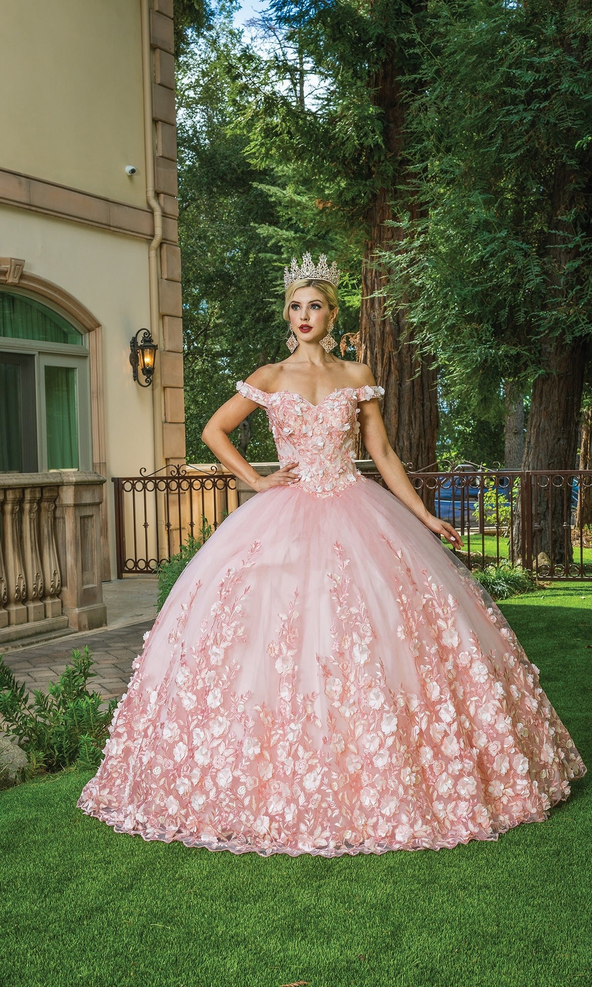 A young woman wearing Quinceanera Dress 1569 by Dancing Queen—an ornate pink ball gown with rose details and off-shoulder straps—stands on grass with a tiara, in front of a building and large trees.