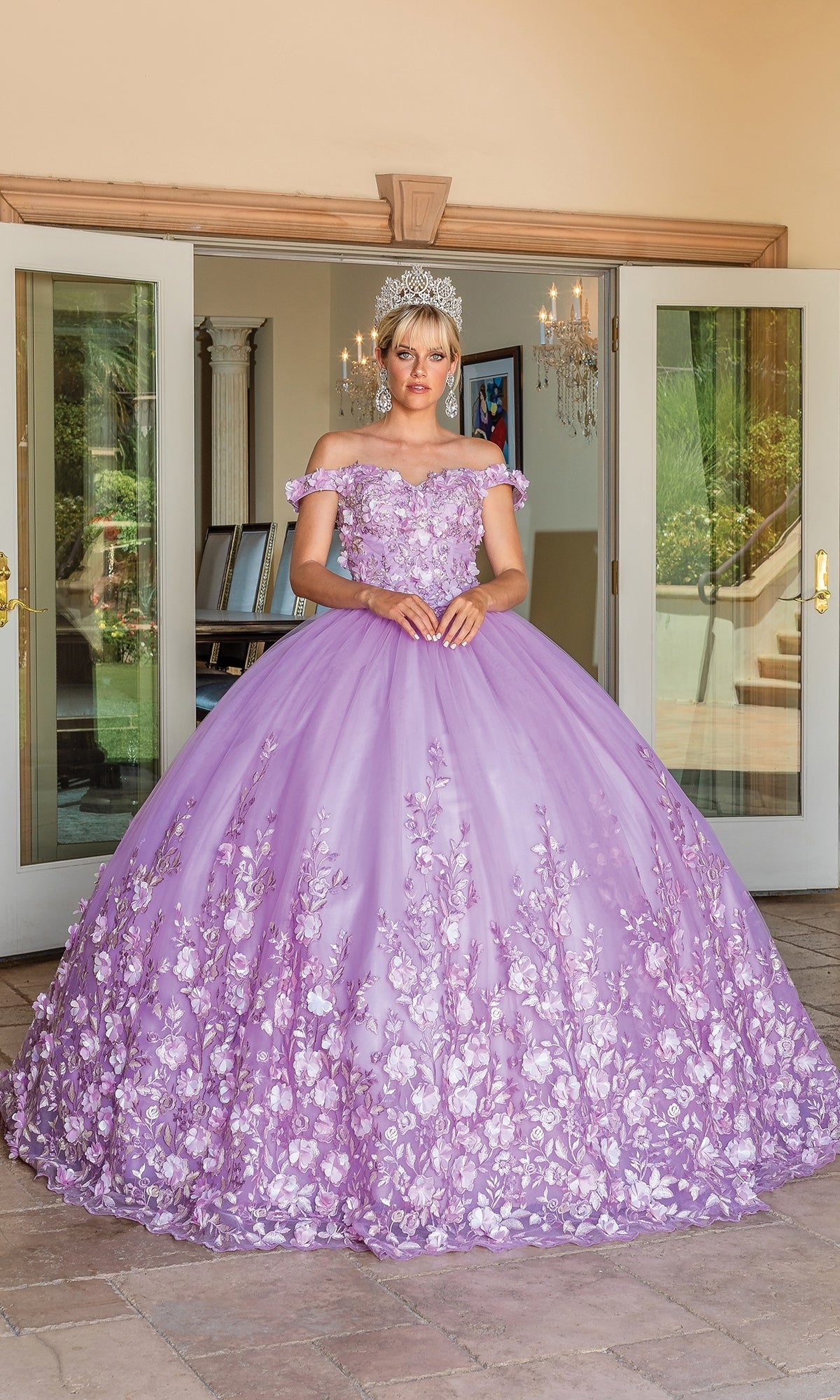 A woman in the Quinceanera Dress 1569 by Dancing Queen, featuring an a-line silhouette, rose details, and off-the-shoulder straps with a tiara, stands in front of open double doors.