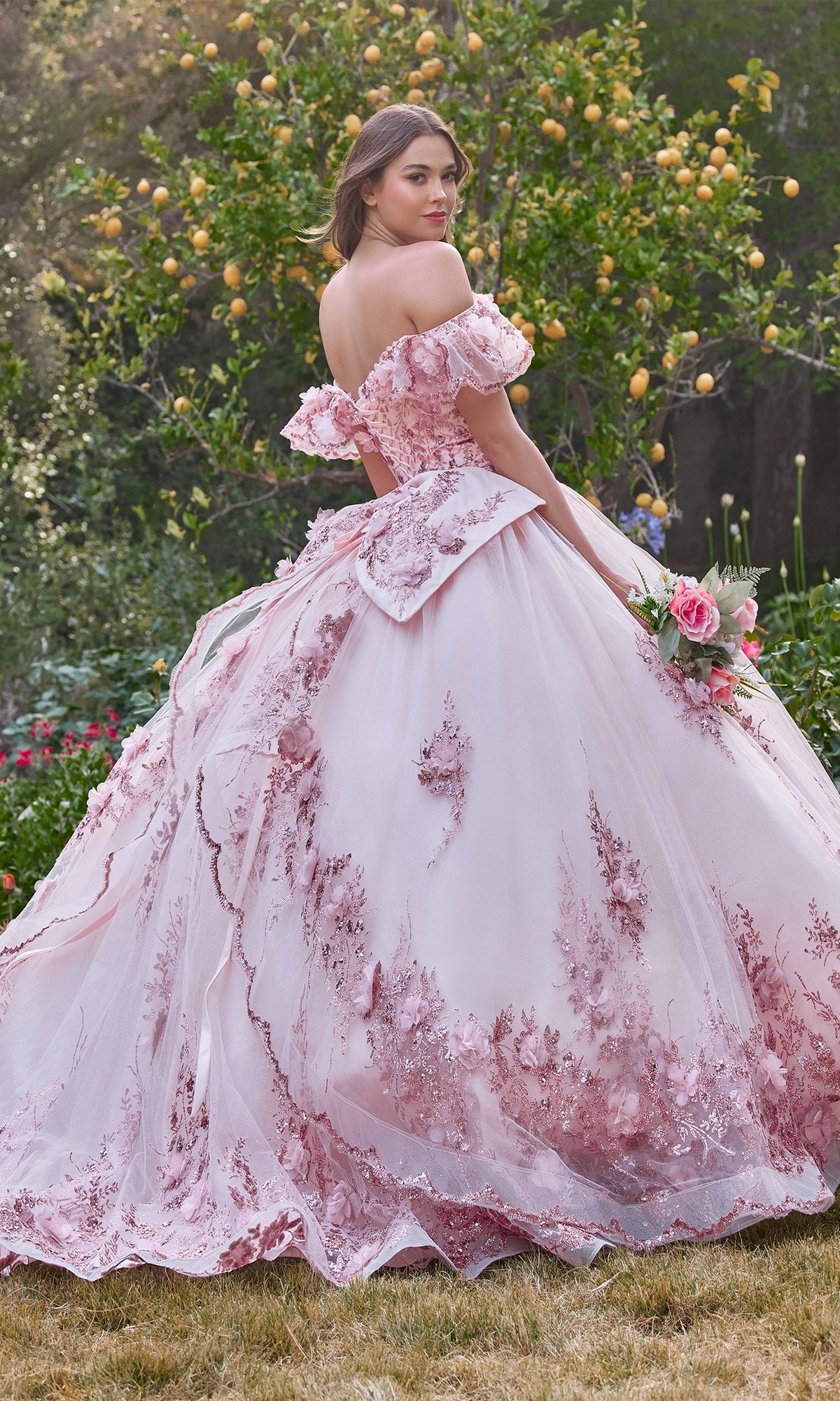 A young woman in a pink Glitter Quince Dress with Back Bow: Ladivine 15701, featuring floral embroidery, stands outdoors with a bouquet among green trees and yellow fruit—the ideal choice for a stunning quinceañera look.