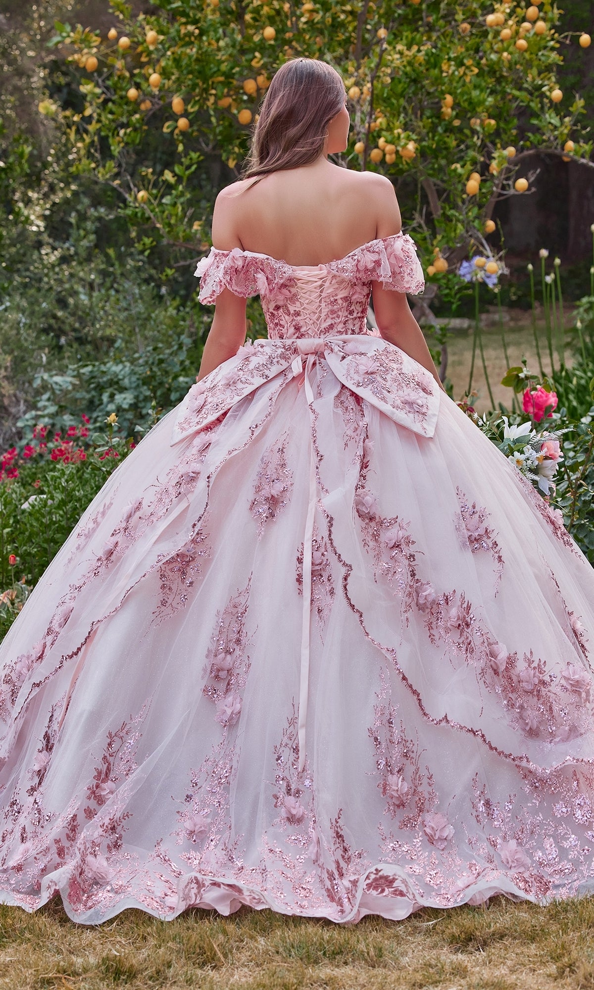 A woman wearing the Glitter Quince Dress with Back Bow: Ladivine 15701, featuring floral embroidery and a large bow, stands in a garden surrounded by lemon trees and blooming flowers.