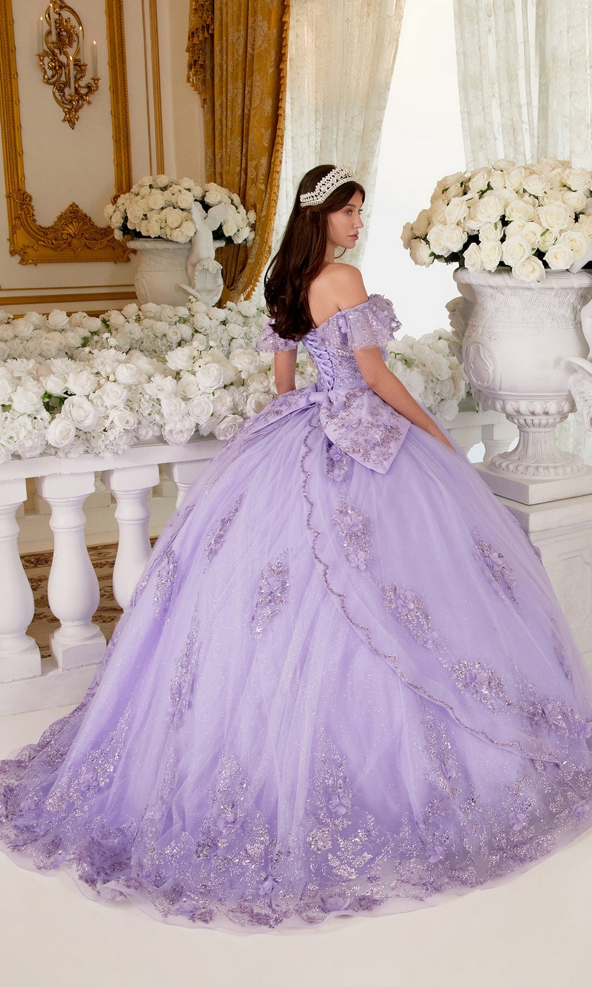 A young woman in the Ladivine 15701 Glitter Quince Dress with back bow, featuring lilac floral details, stands by white columns and large white flower arrangements, facing slightly away from the camera.