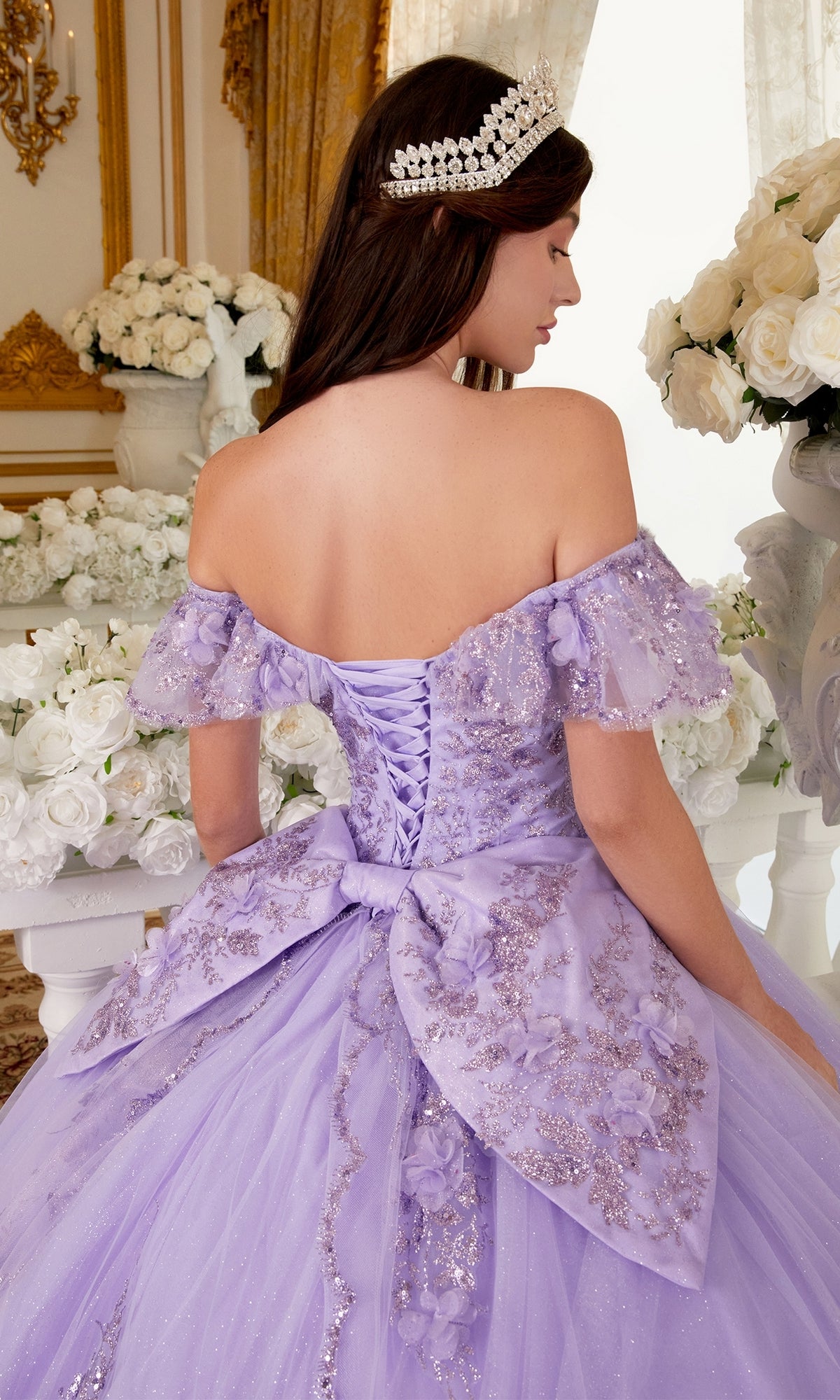 Wearing the Ladivine 15701 Glitter Quince Dress with a back bow and off-the-shoulder sleeves, a woman with long dark hair and a pearl headpiece stands among white roses, facing away.