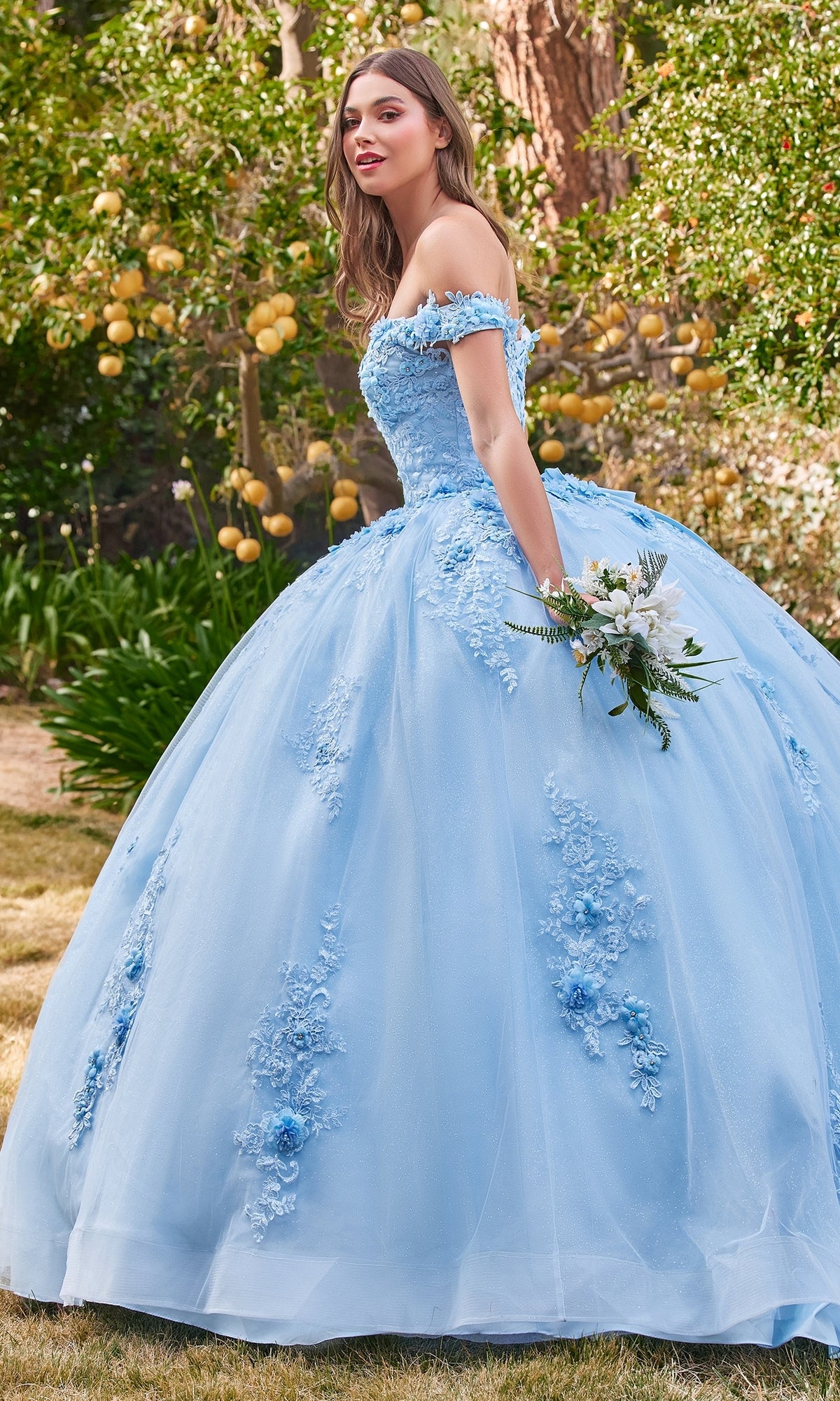 A woman stands in a garden holding a bouquet, wearing the Ladivine 15702 quinceañera dress featuring light blue fabric, off-the-shoulder styling, and 3-D floral details.