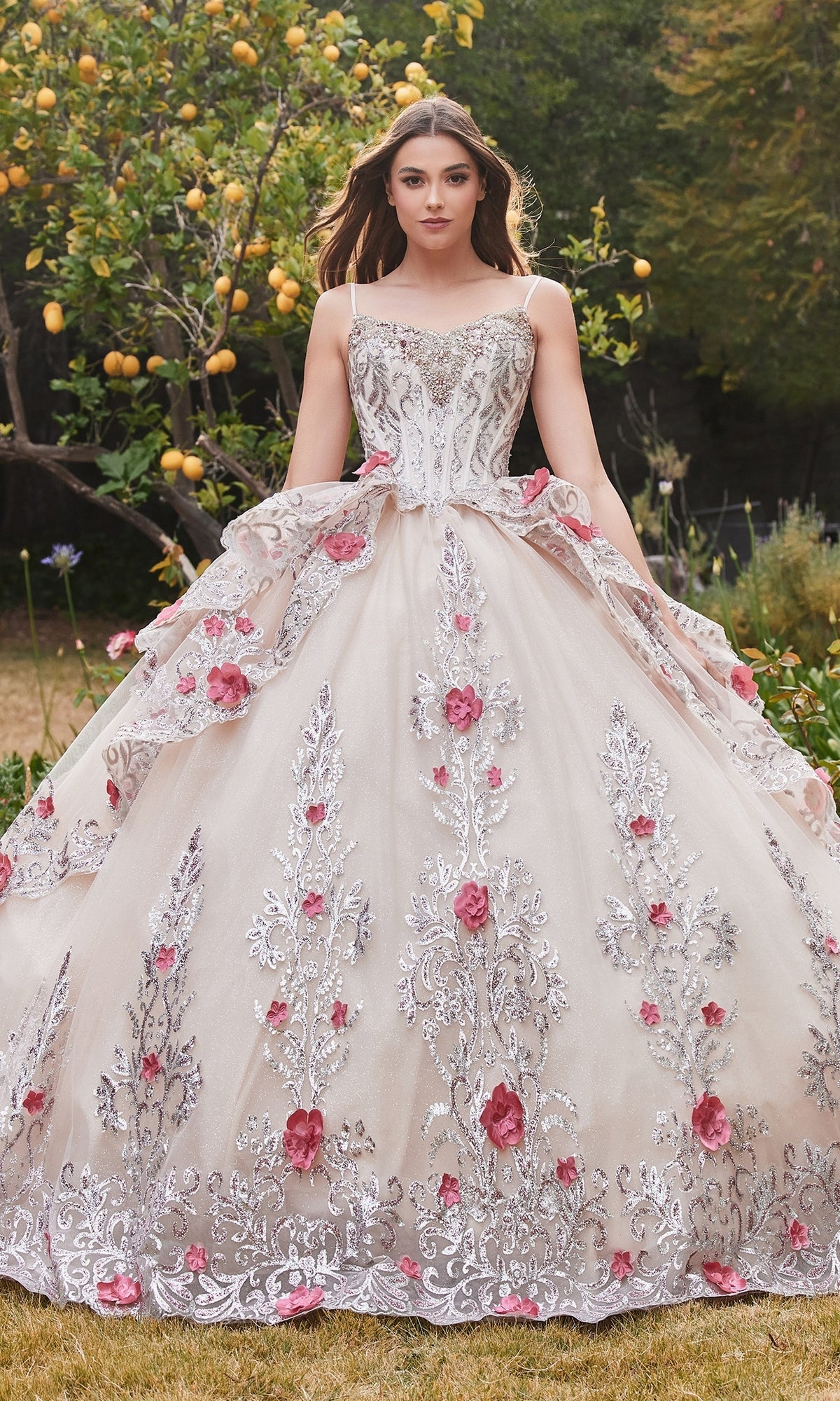 A woman stands outdoors in the Ladivine 15703 Glitter Champagne Quinceañera Dress, featuring silver embroidery and pink floral accents, surrounded by lush greenery and a lemon tree.