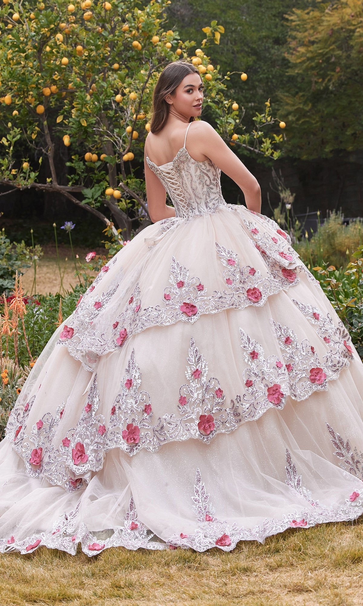 A young woman outdoors wears the Ladivine 15703 Glitter Champagne Quinceañera Dress, featuring floral embroidery and pink accents, as she looks over her shoulder.