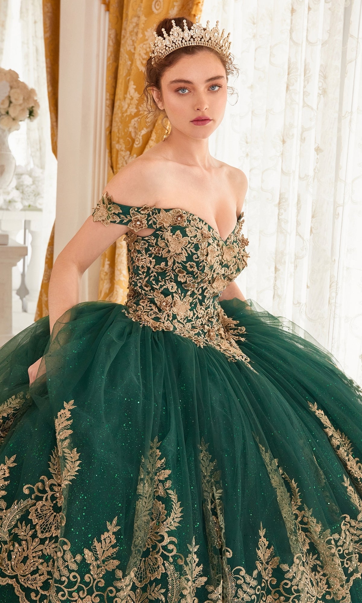 A woman wearing the Ladivine 15705 Quince Dress with gold embroidery and a tiara stands indoors before sheer curtains and gold drapes.