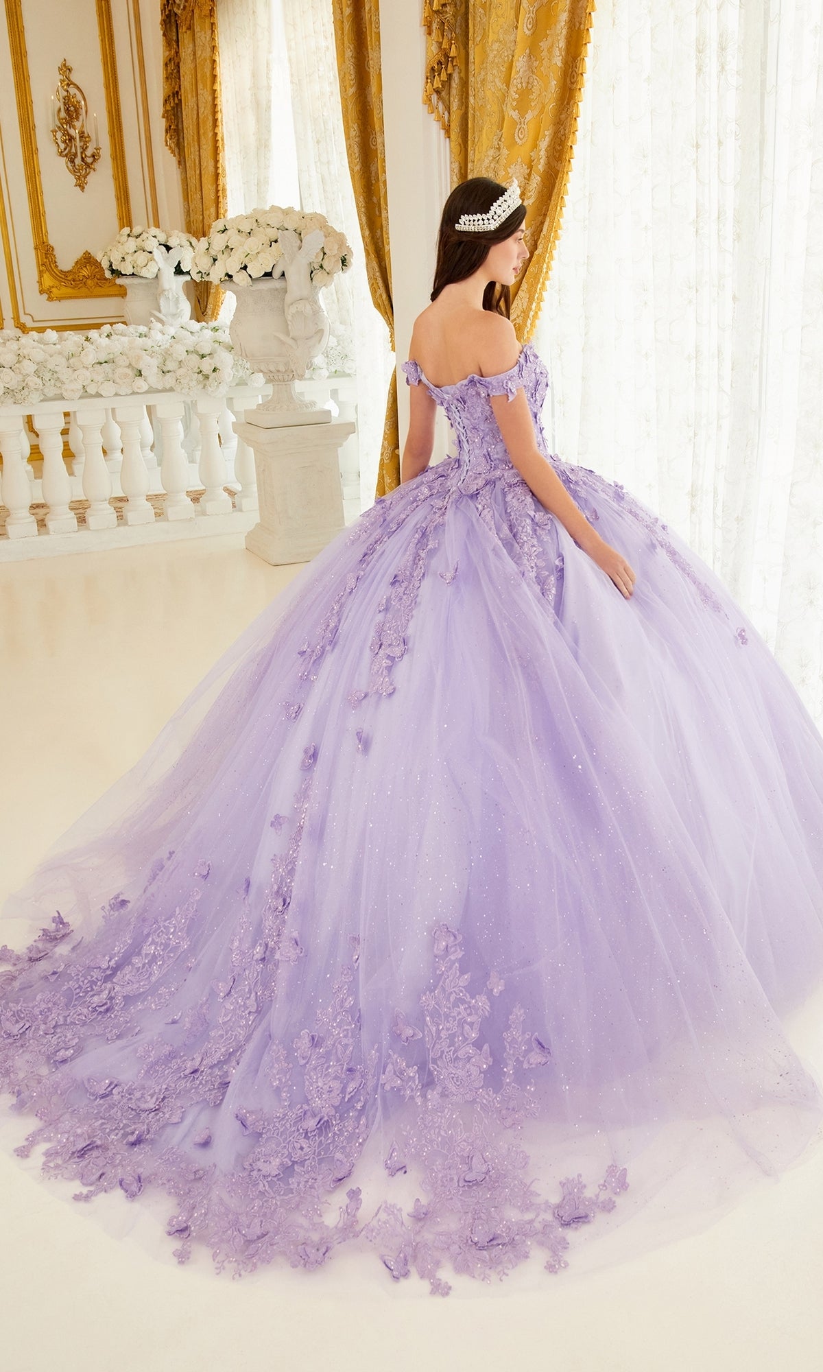 A woman in the Ladivine 15709 Lavender Purple Quinceañera Dress with floral details and a tiara stands in an ornate, gold-accented room featuring large windows and white flowers.