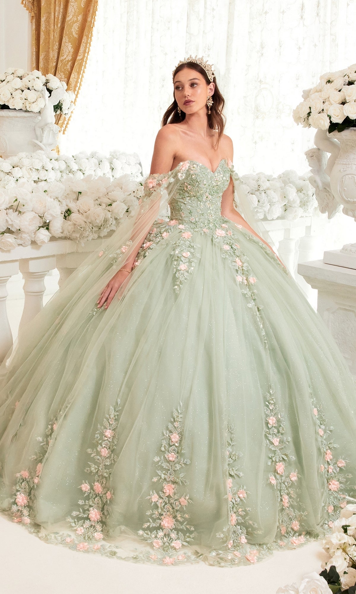 A woman in the Ladivine 15716 Sage Green Quinceañera Dress with floral embroidery stands in an elegant room adorned with white flowers and gold curtains.