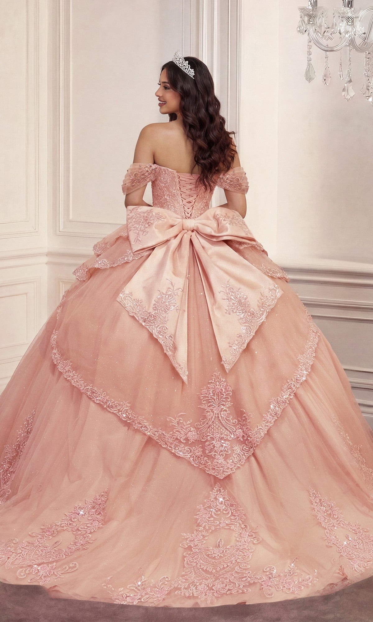 Wearing the Quinceanera Dress 15729 by Ladivine, a woman stands facing away in an ornate room, showcasing the pink off-the-shoulder ballgown embellished with intricate floral lace appliques and a large bow.
