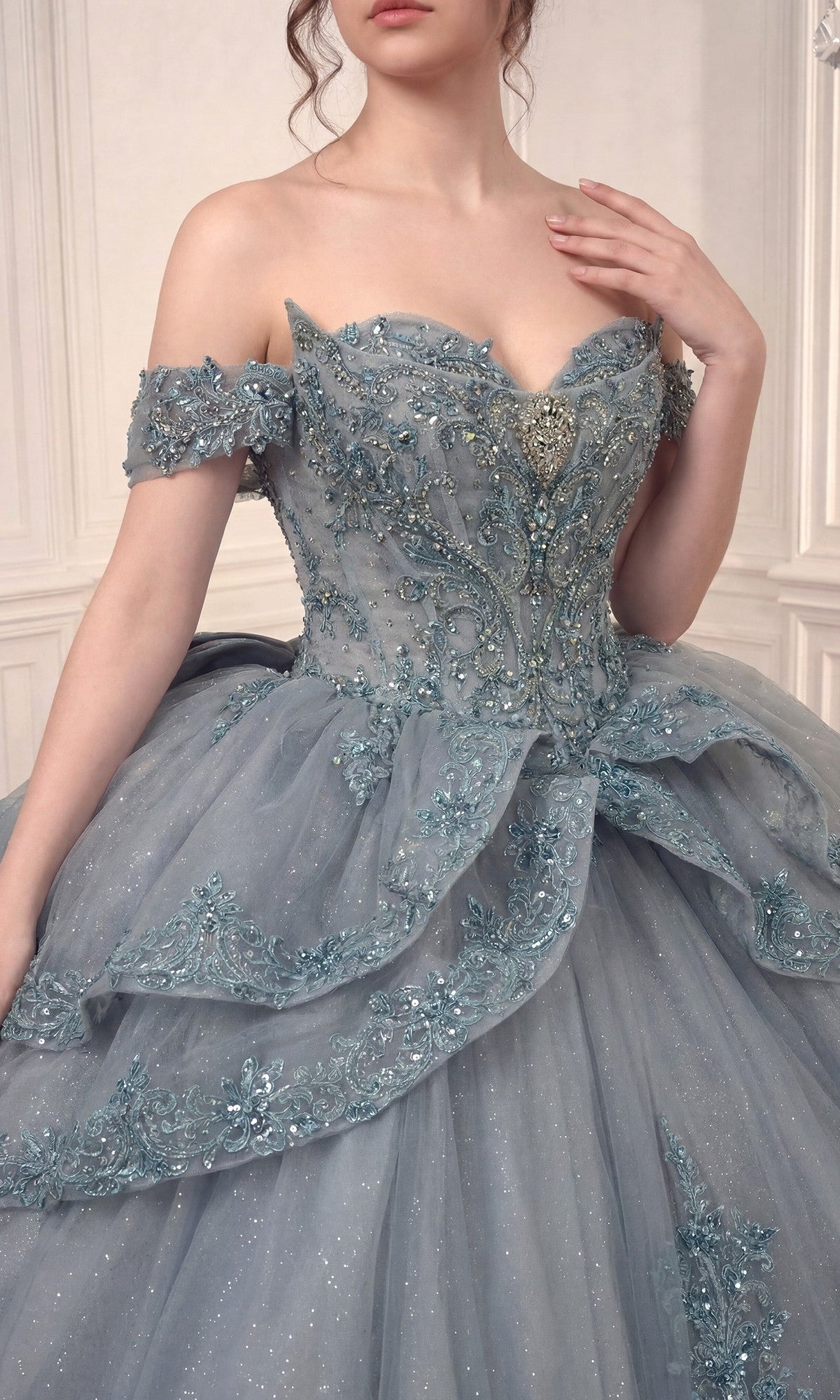 A woman wearing the Quinceanera Dress 15729 by Ladivine, an off-the-shoulder ballgown with a layered tulle skirt and intricate floral lace embroidery and appliques, standing indoors.