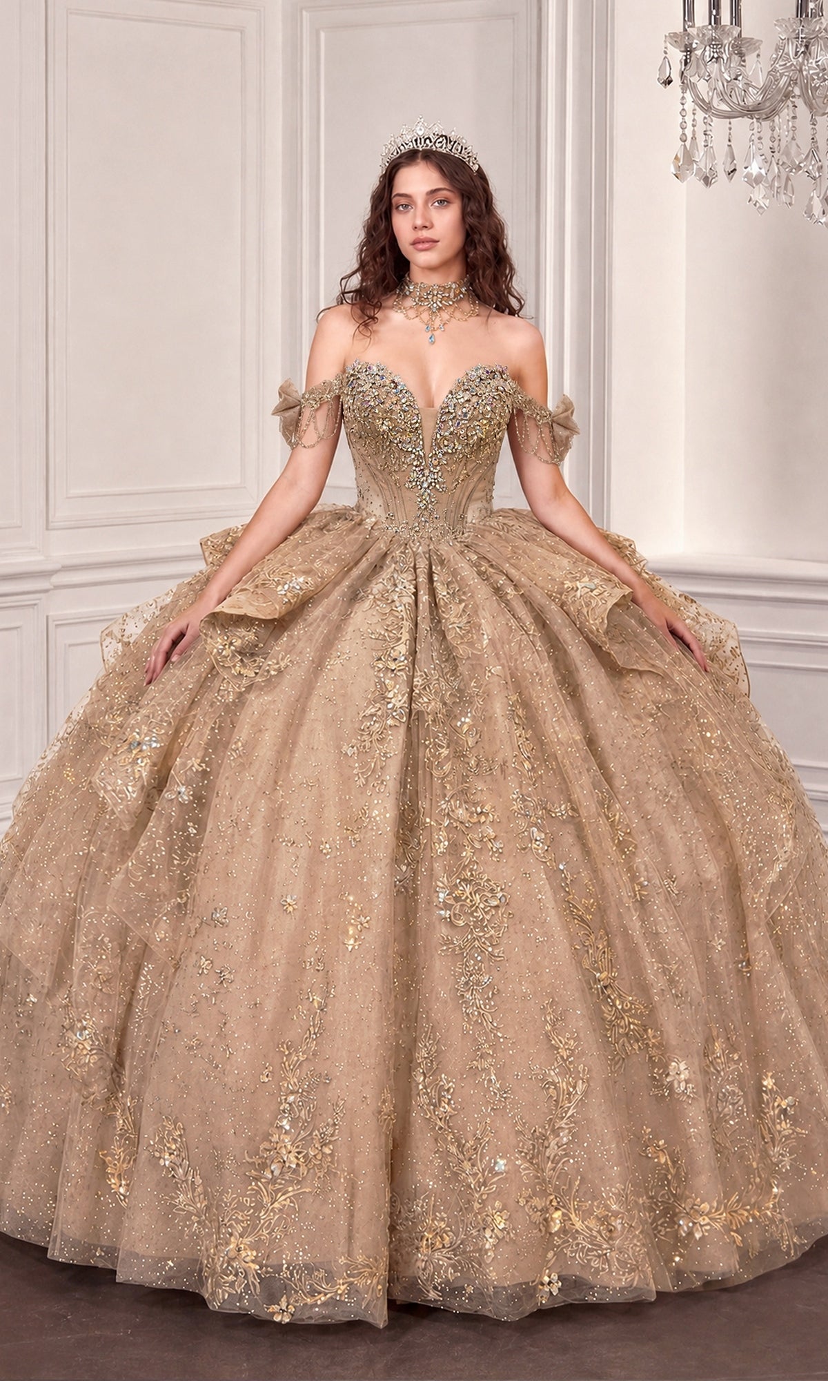 A woman stands indoors against a white paneled wall, illuminated by the chandelier overhead, wearing the Quinceanera Dress 15743 By Ladivine in Mocha Gold. The ornate off-the-shoulder gown features intricate embroidery and shimmers with elegant beaded details, while a sparkling tiara completes her opulent look.