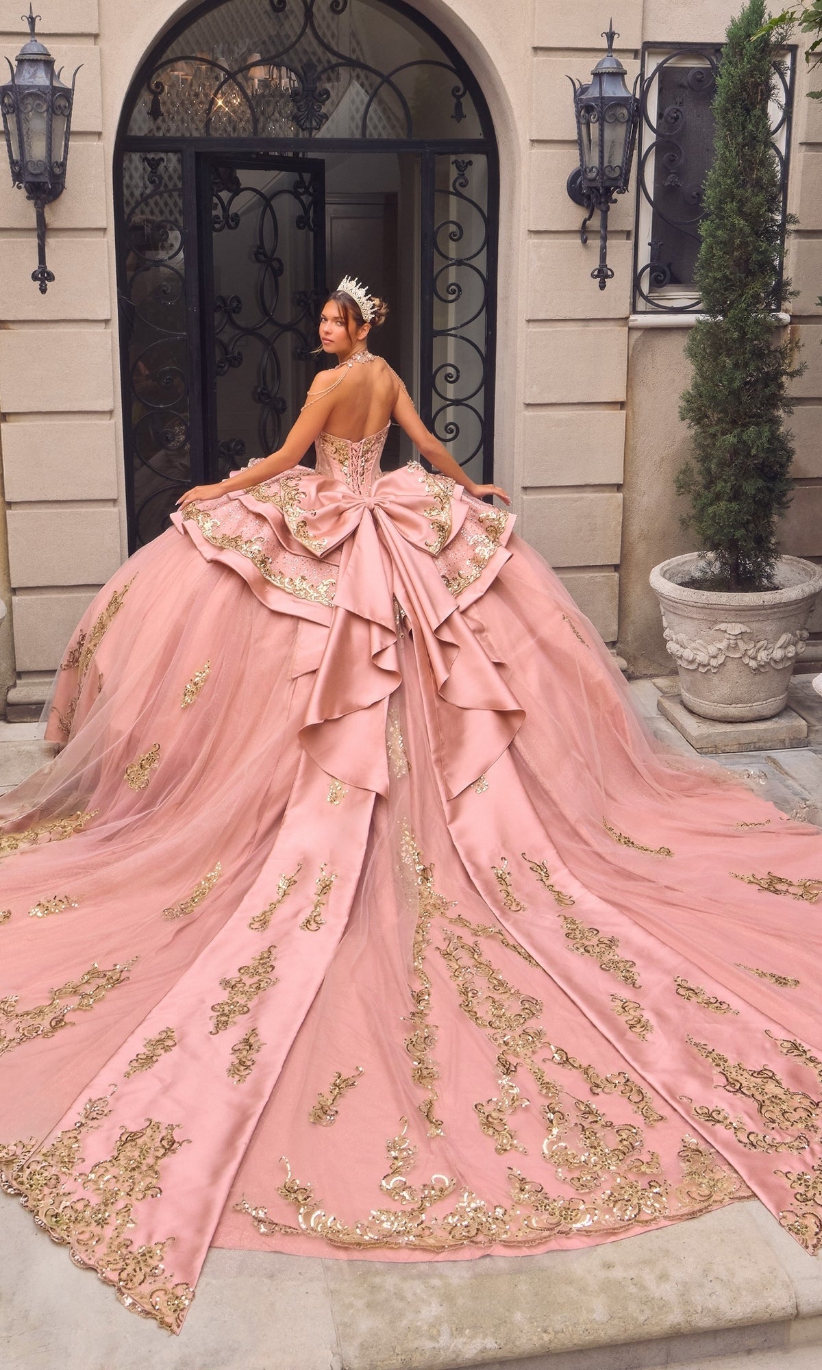 A woman stands in front of an ornate doorway, facing away from the camera, wearing the Quinceanera Dress 15744 by Ladivine—a pink and gold embellished glitter tulle ballgown with a long train.