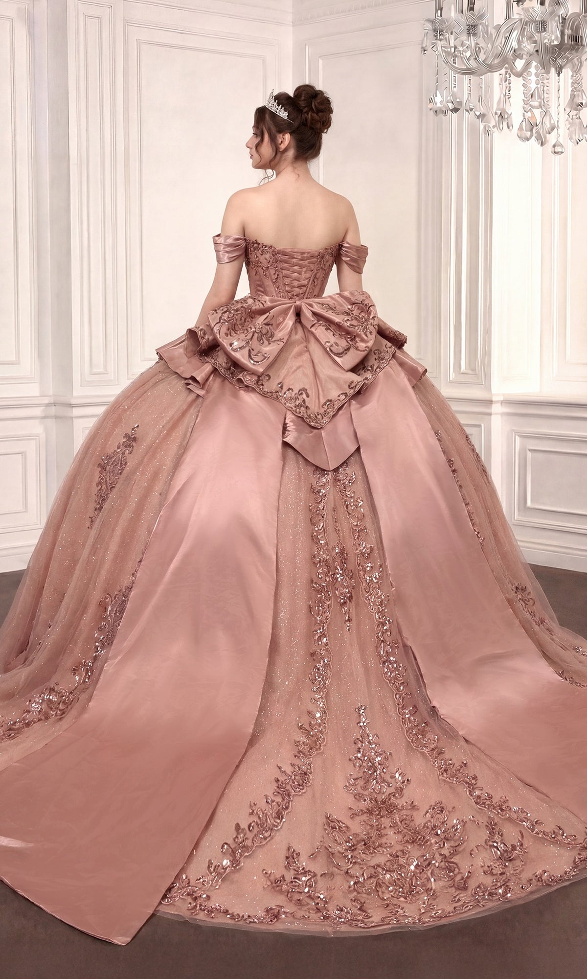 A woman stands facing away in an ornate room, wearing the Quinceanera Dress 15745 By Ladivine—a pink, off-the-shoulder ballgown featuring a beaded corset bodice and lace details.