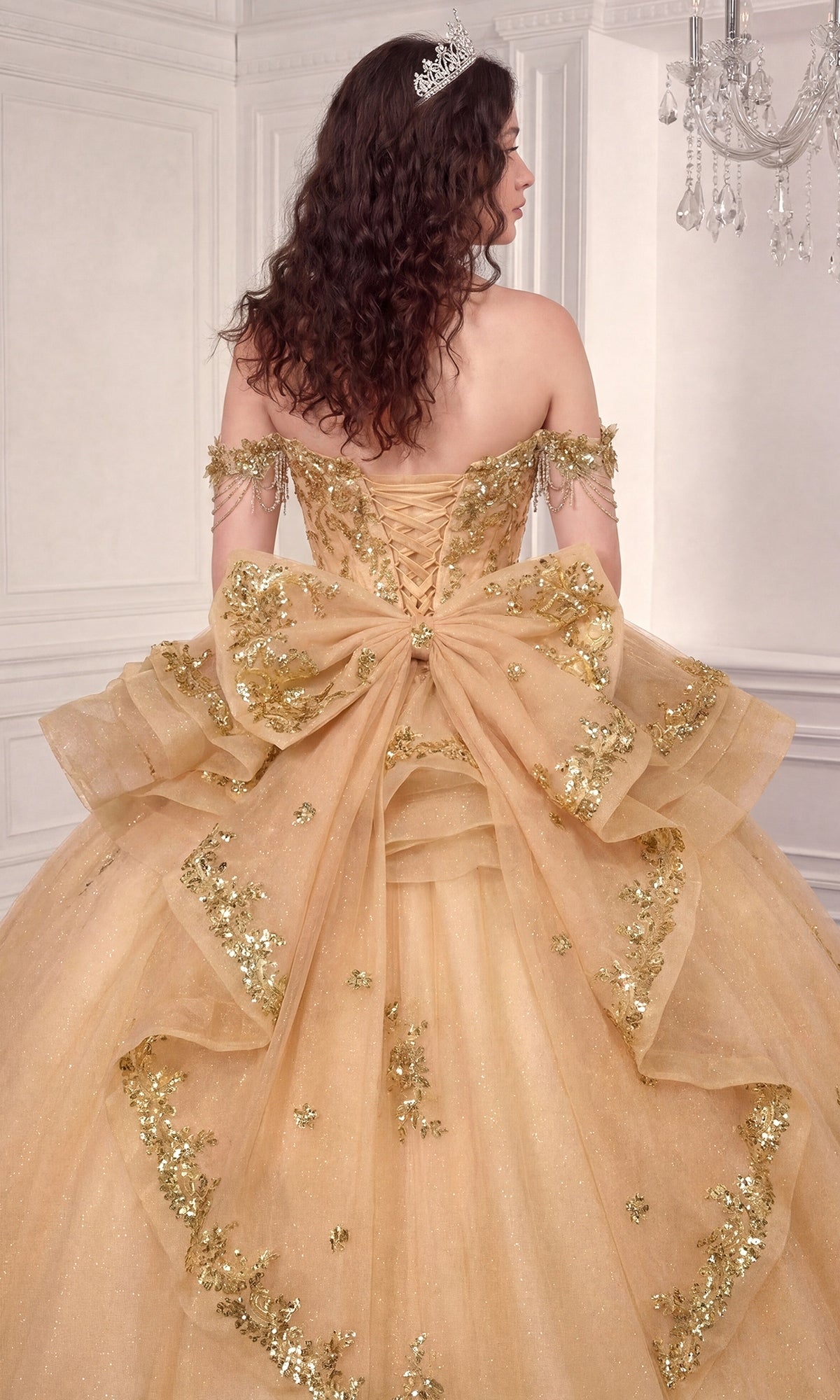 A woman in a gold, off-the-shoulder ballgown adorned with sequins and a large bow stands facing away in an elegant white room, showcasing the stunning silhouette of the Quinceanera Dress 15746 by Ladivine.